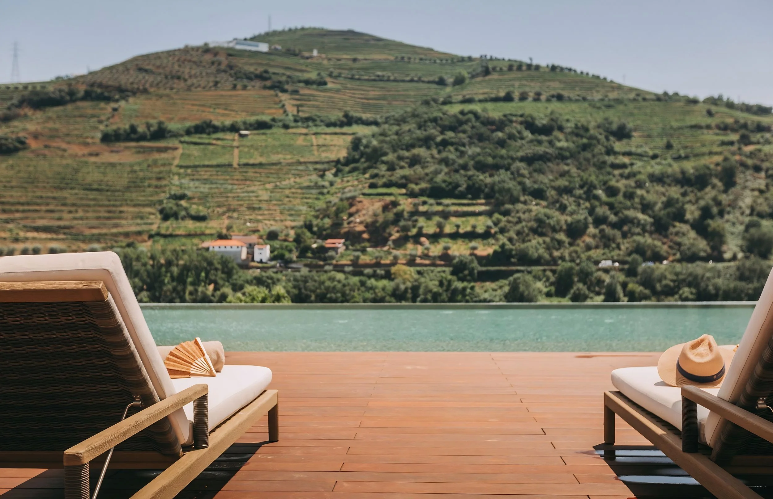Poolside view with two lounge chairs, a sunhat, and a folding fan on a wooden deck. In the background, there are green hills with terraced vineyards and a clear sky.