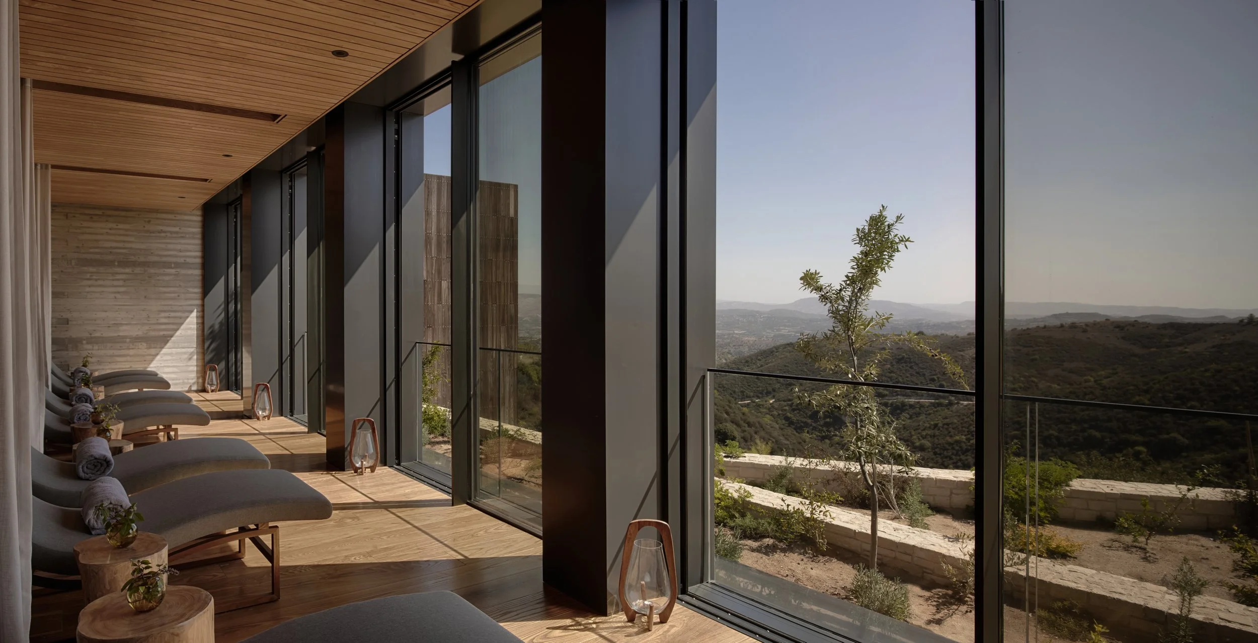 Interior of a modern room with floor-to-ceiling windows showing a landscape of rolling hills and a clear sky. The room has wooden floors, a wooden ceiling, and lounge chairs with rolled towels and small plants.