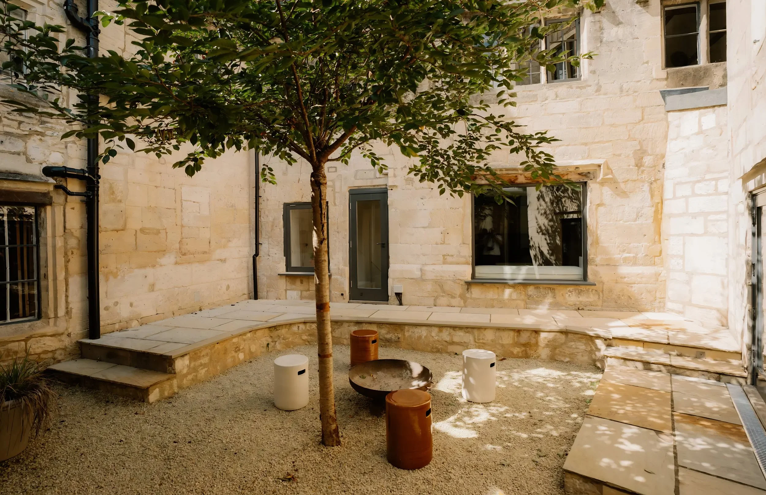 Small enclosed courtyard with a young tree at the center, surrounded by a cream-colored stone building. There are windows with gray frames, a glass door, and wooden steps leading to the door. Decorative cylindrical planters and tiles are placed aroun