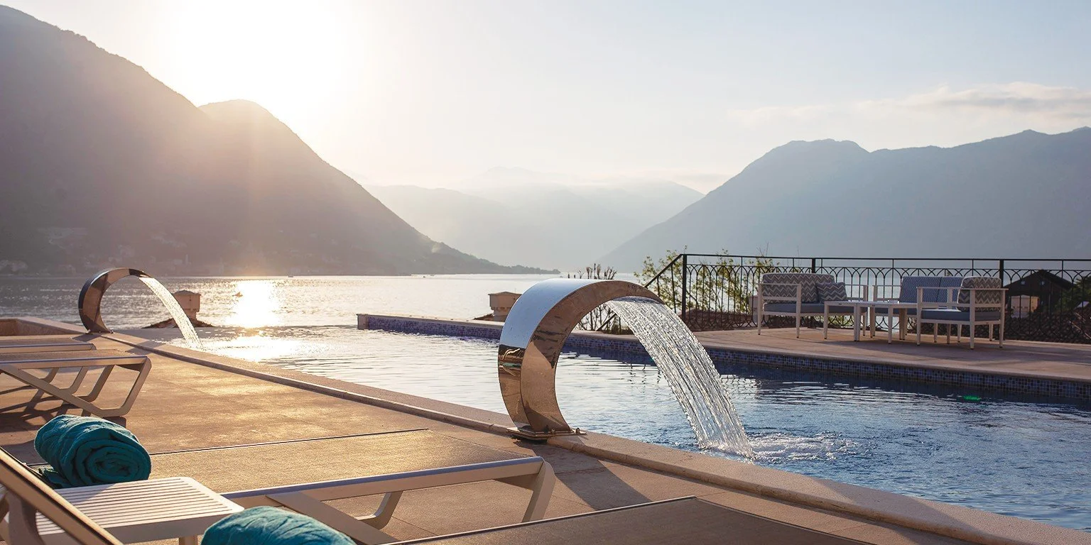 A scenic outdoor pool area with flowing water features, lounge chairs, and a mountain view during sunset or sunrise.