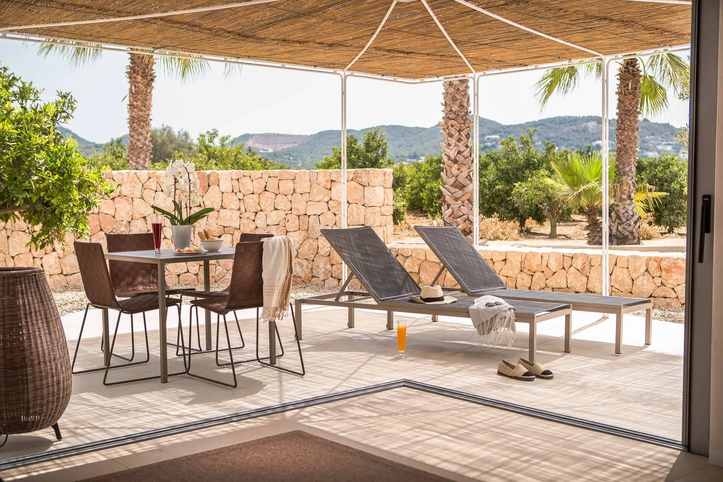 An outdoor patio area with lounge chairs, a table with drinks, potted plants, and a stone wall, with mountains and trees in the background.