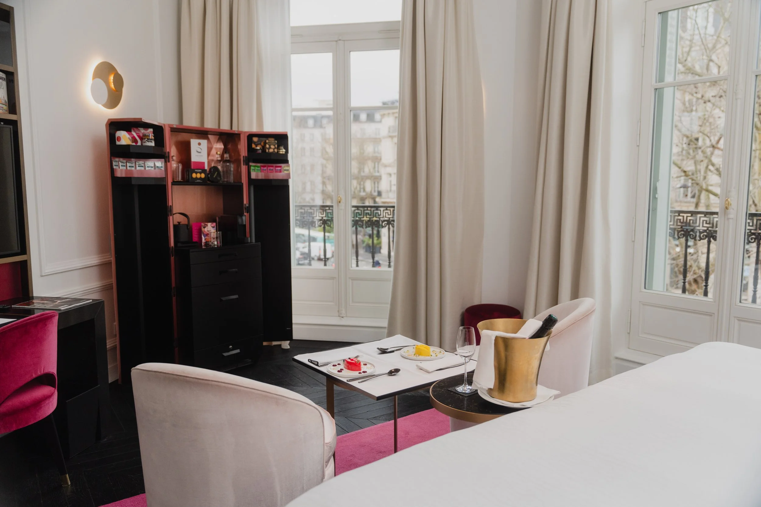 Elegant dining room with large windows, cream-colored curtains, and a mix of modern and vintage furniture. A small table with a place setting and a glass of water is visible, with some plates of food and utensils. A black cabinet with glass jars and pink packaging is in the corner, and a decorative wall lamp is on the wall.