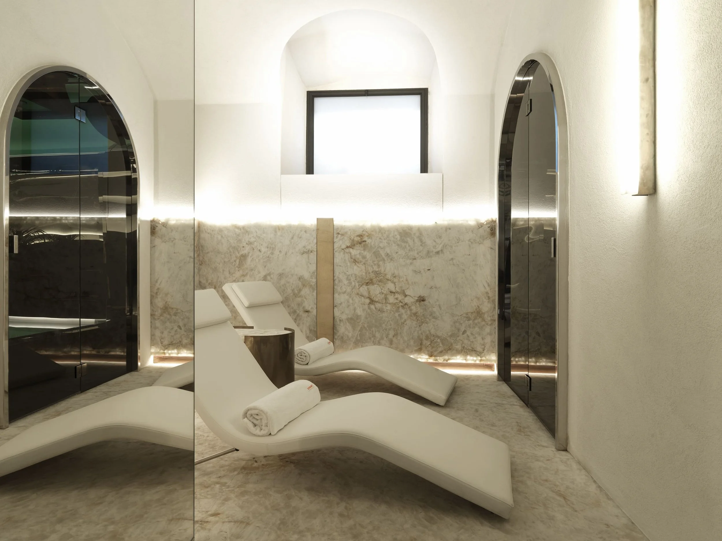 Modern spa room with white lounge chairs, white towels, marble accents, and curved mirrors. Bright lighting and minimalist decor.