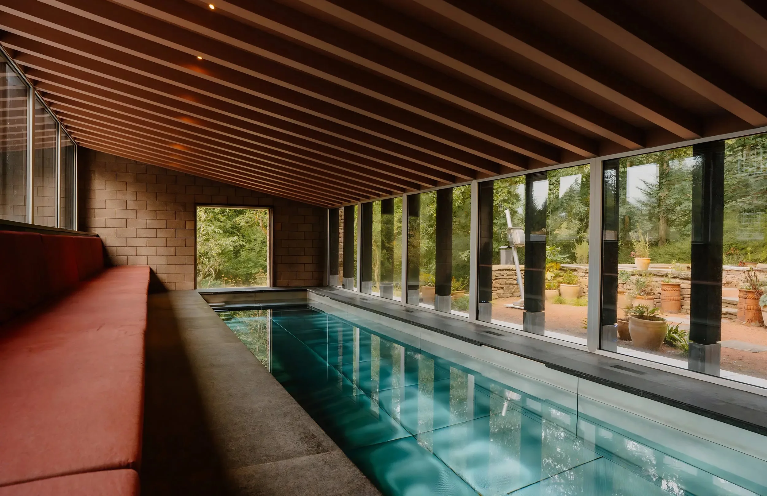 Indoor pool area with large glass windows showing a garden outside, a brick wall, and a wooden ceiling.