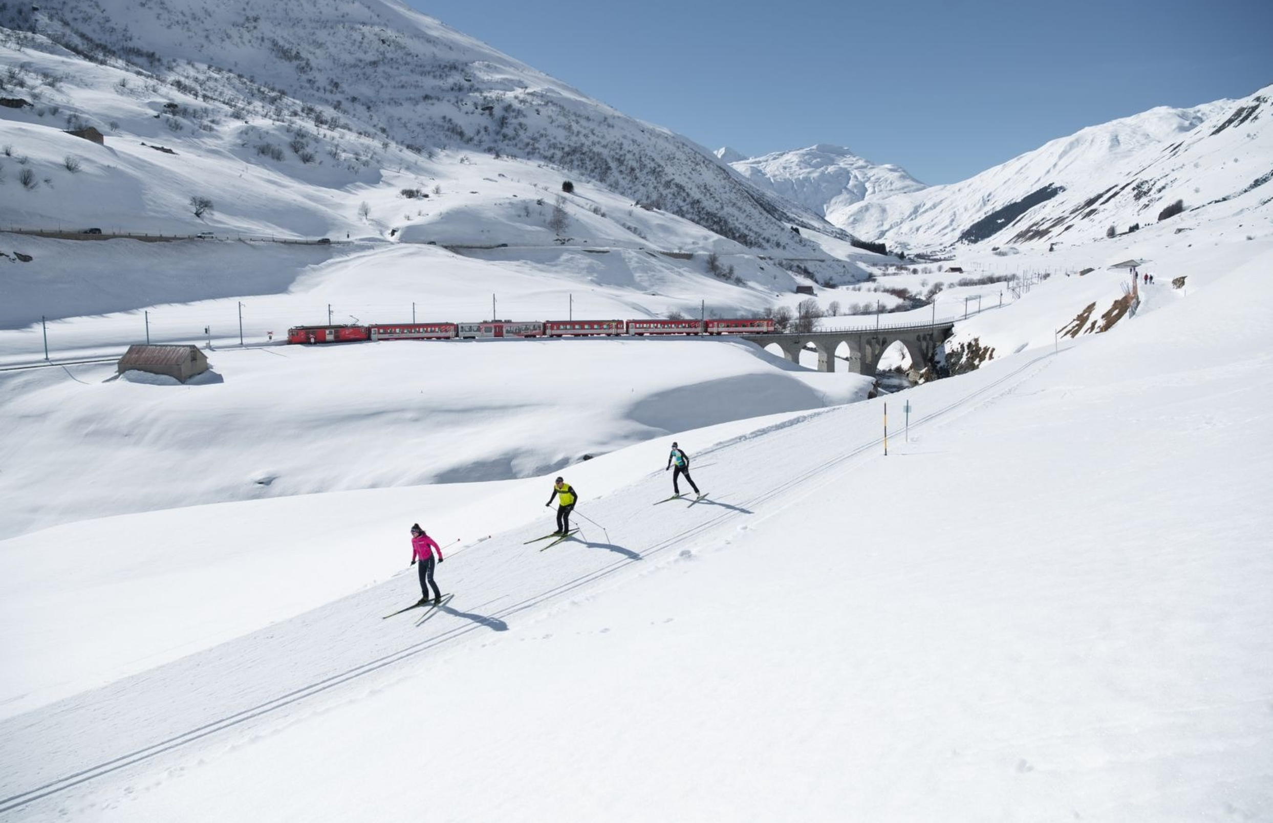 A snowy mountain landscape with a red train crossing a bridge, and ski tourists skiing on snow-covered slopes.