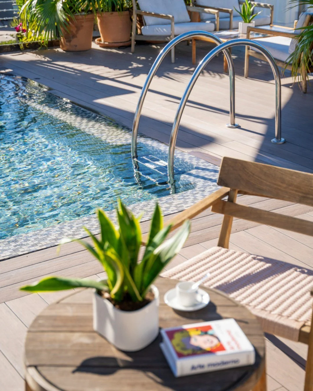 An outdoor pool area with wooden deck, metal pool ladder, potted plants, wooden lounge chairs, and a table with a potted plant, a cup and saucer, and a book.