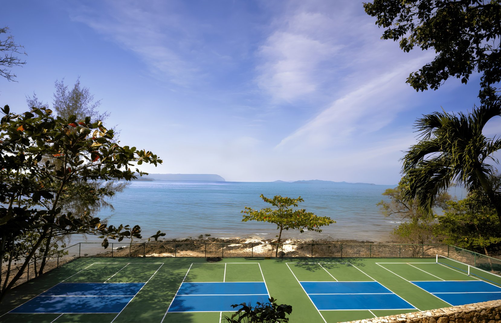 Tennis courts overlooking the ocean at a tropical beach, with trees and a rocky shoreline in the foreground, and a clear blue sky with a few clouds.