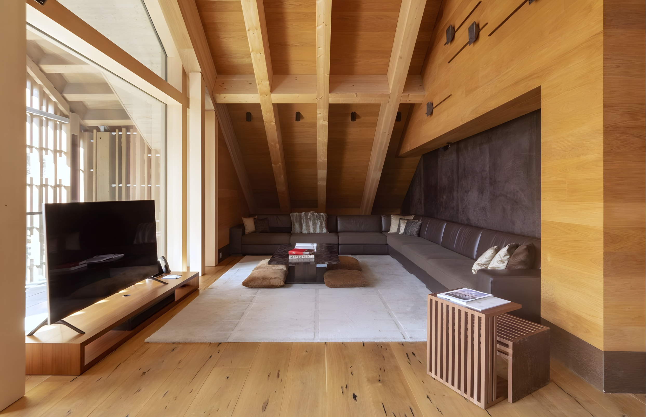 Living room with wooden ceiling beams, a large sectional sofa, a coffee table with books and pillows, a TV on a wooden stand, and large windows with blinds.