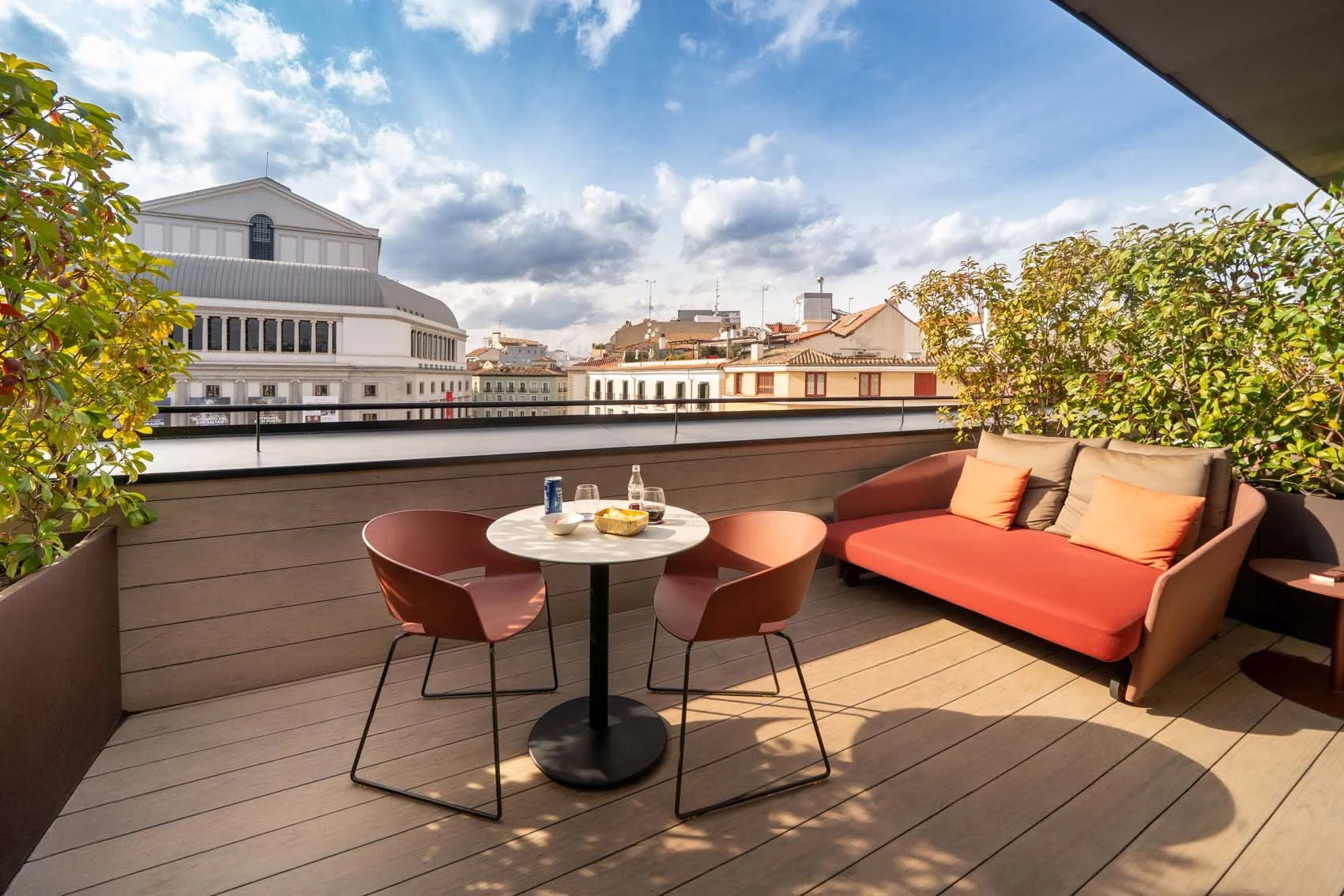 A rooftop patio with a round table, two chairs, and a sofa with cushions. There are drinks and snacks on the table. The patio has potted plants and overlooks a city skyline with buildings and a partly cloudy sky.