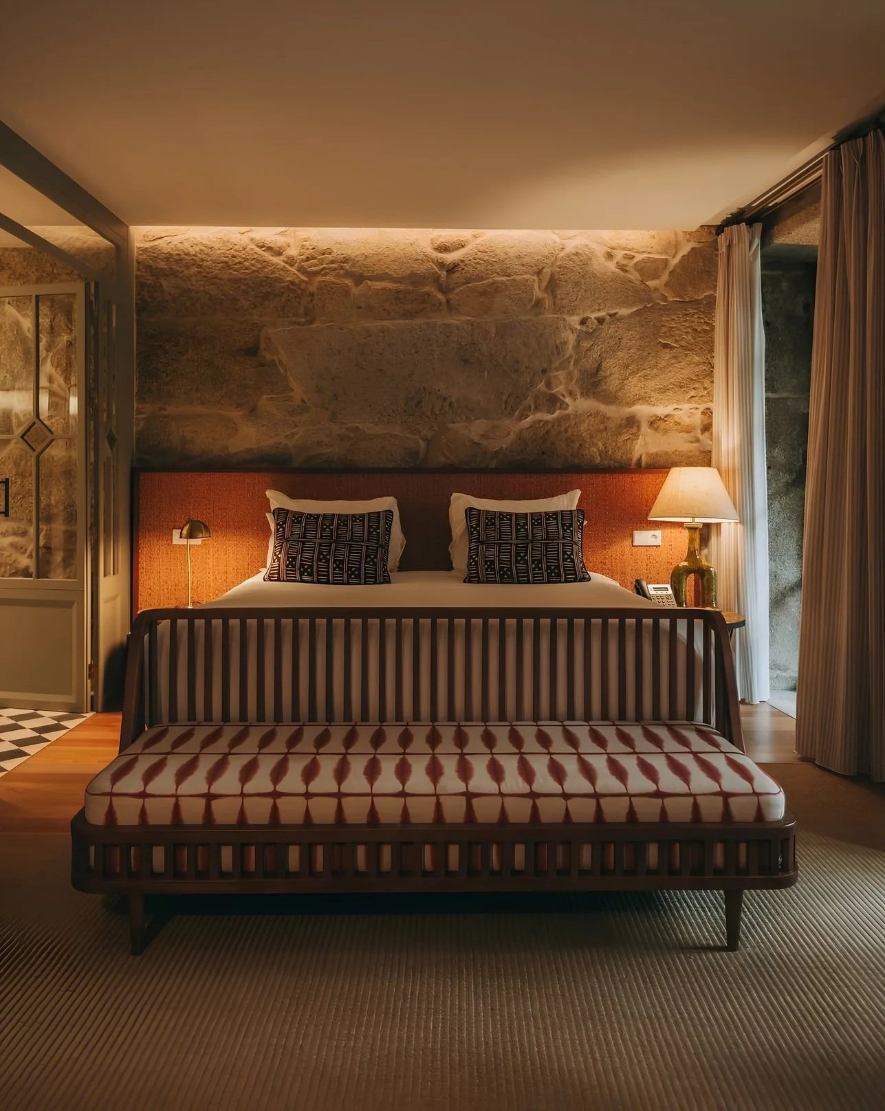 A hotel room with a large bed, decorative pillows, a wooden bed frame, and a stone accent wall. There is a bench at the foot of the bed, a side table with a lamp and a telephone, and curtains covering a window or balcony door.
