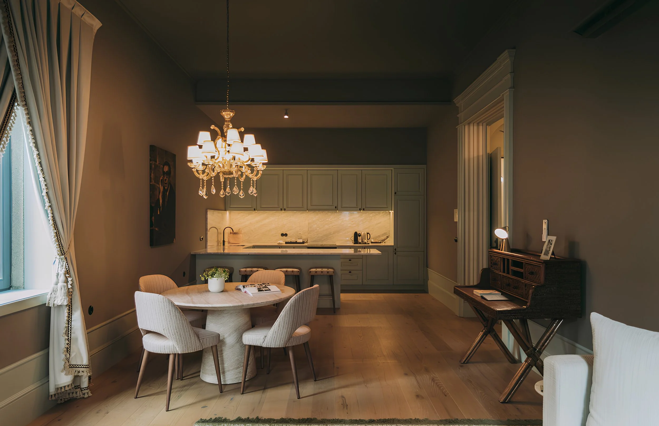 A cozy dining and kitchen area with a chandelier, round dining table with white chairs, and a kitchen with light gray cabinets, marble backsplash, and wooden flooring.