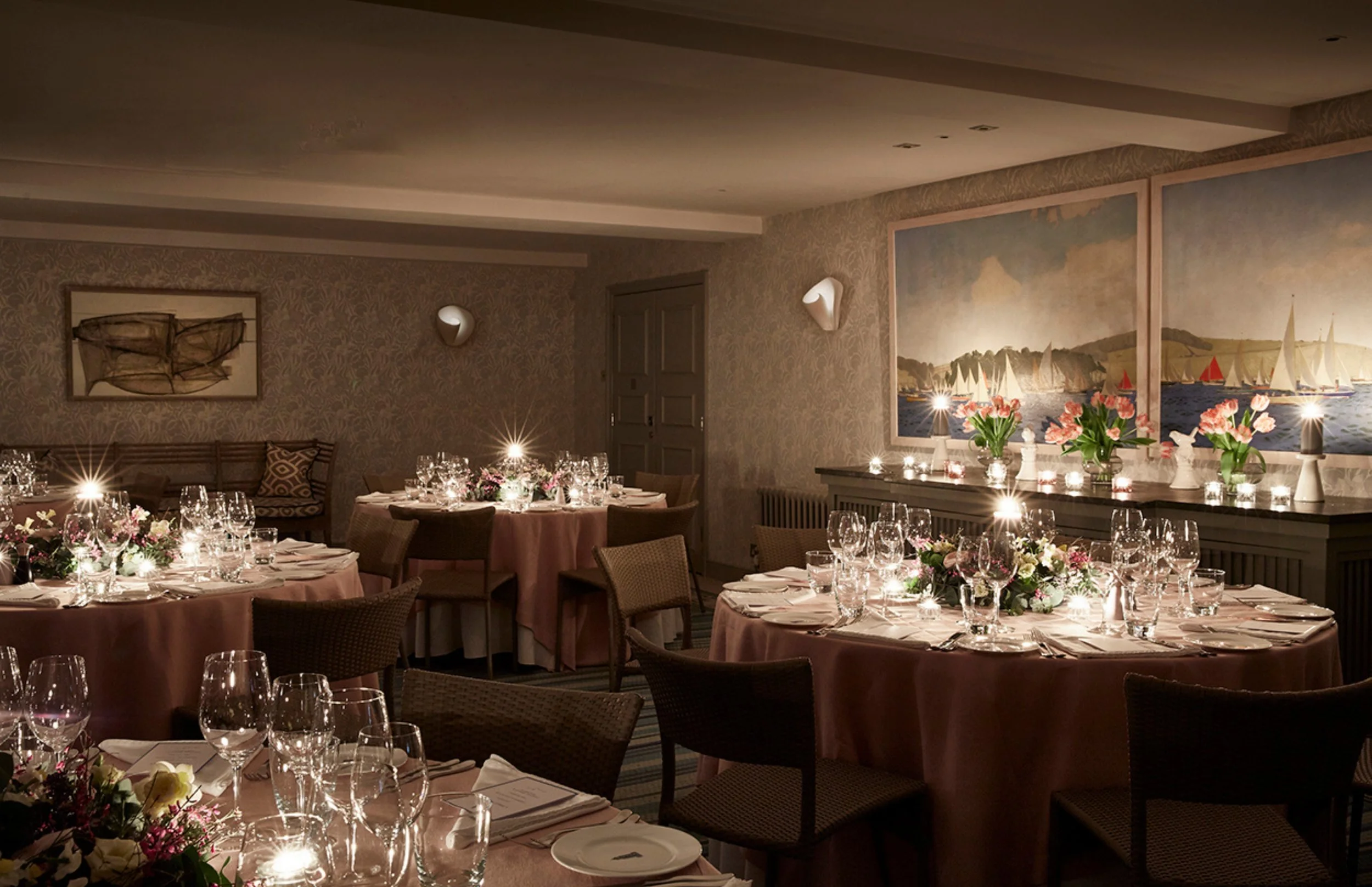 A formal dining room set for a banquet with round tables covered in pink tablecloths, floral centerpieces, and multiple wine glasses. The room has walls with patterned wallpaper, framed artwork, and a large painting of sailboats on water.