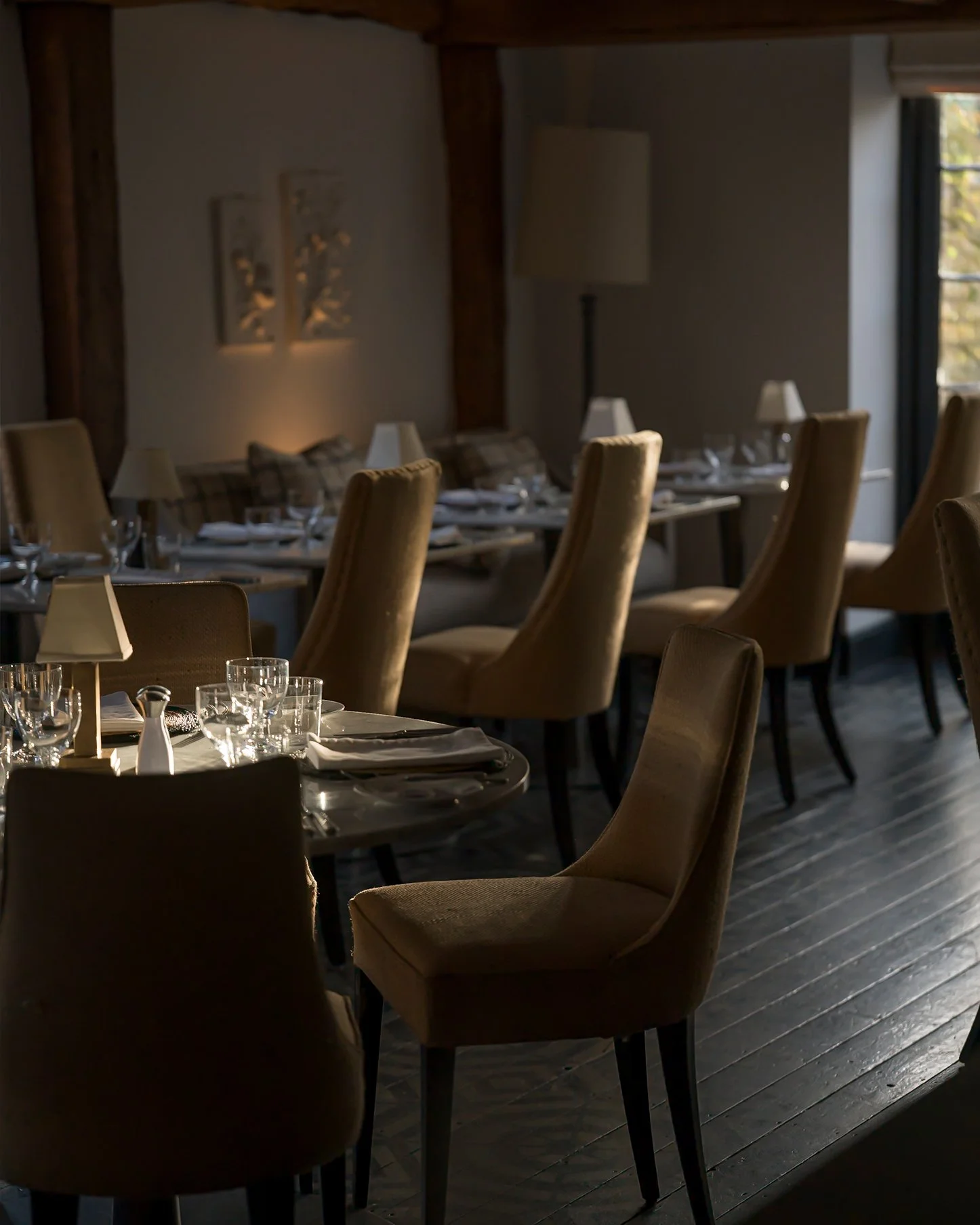 Empty dining room with tables set with glasses, plates, napkins, and small lamps, beige upholstered chairs, and natural light from a window.
