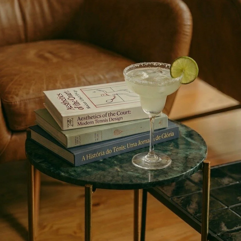 A martini glass with salt rim and a lime wedge on the rim, placed on a small round marble table with a stack of three books underneath. The setting appears cozy with a wooden floor and brown leather chair in the background.