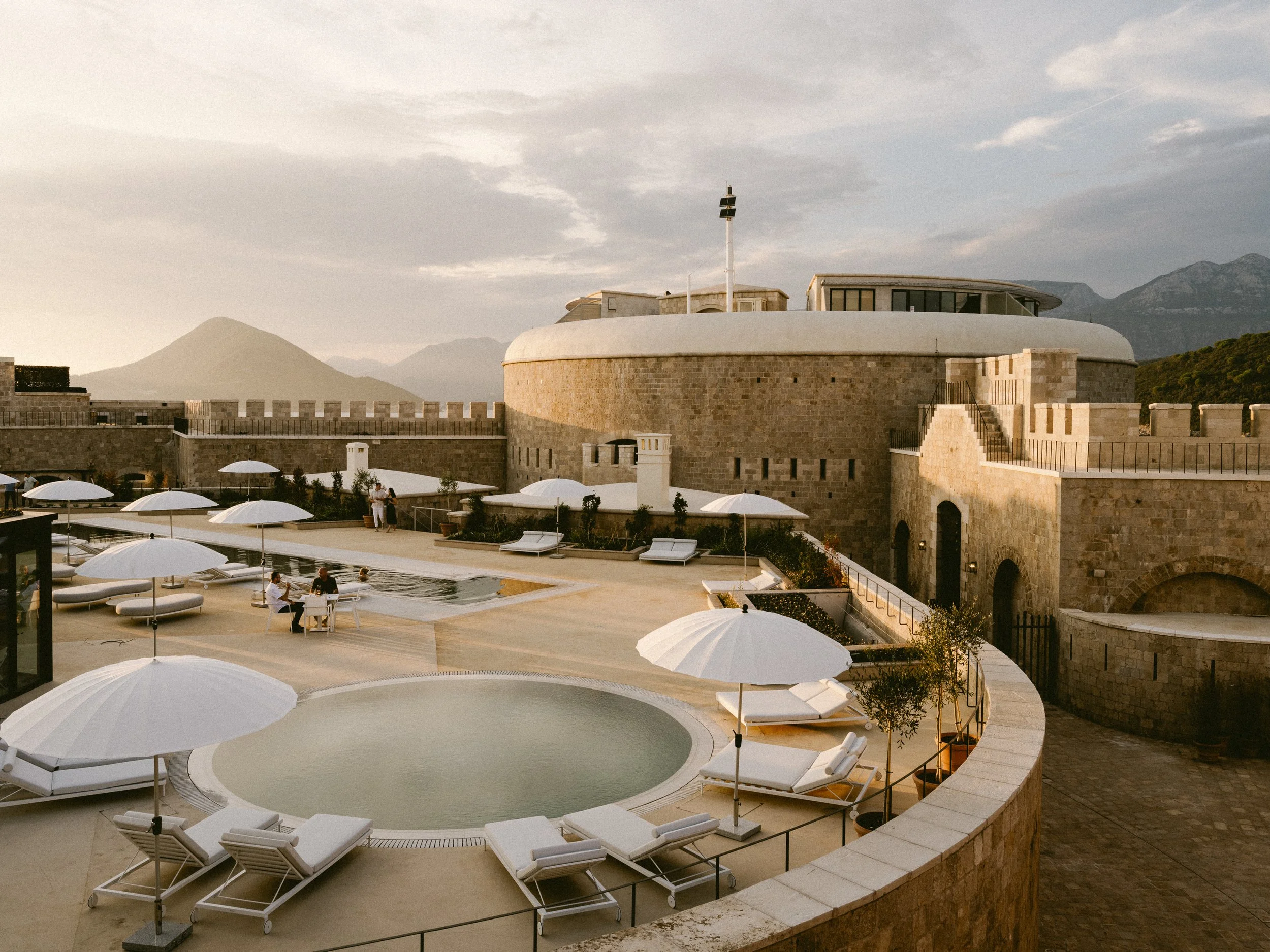 A luxurious outdoor swimming pool area with white umbrellas, lounge chairs, and a hot tub. In the background, stone buildings with a castle-like appearance and a mountainous landscape under a cloudy sky.