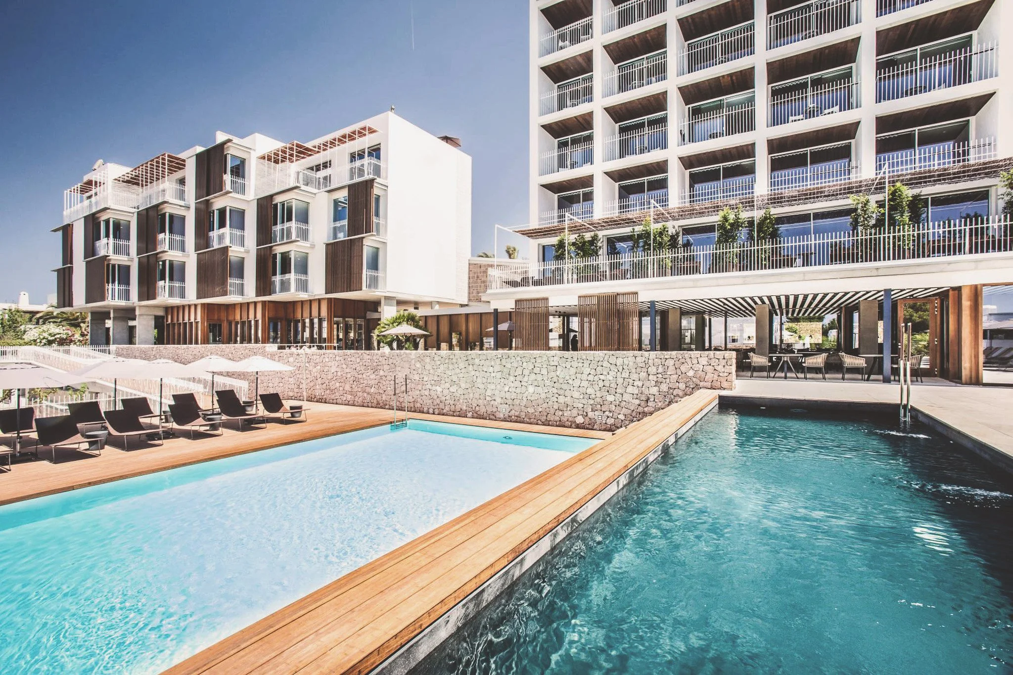Modern apartment building with balconies overlooking a swimming pool area, lounge chairs, and outdoor seating under umbrellas, with clear blue sky.