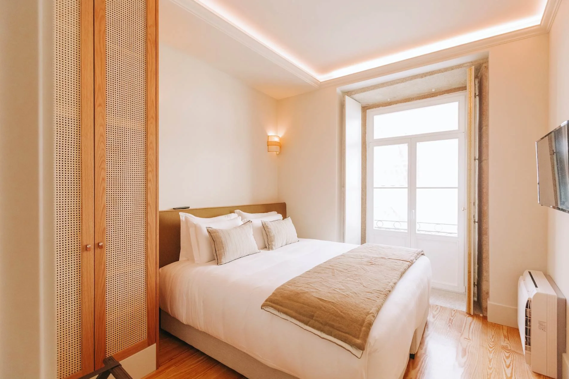 A cozy hotel bedroom with a large bed made with white linens, beige pillows, and a beige bed runner. There's a wooden wardrobe with a lattice design on the left and a large window with white framing allowing natural light. A wall-mounted light fixture is on the beige wall beside the bed, and the ceiling has a cove lighting feature.