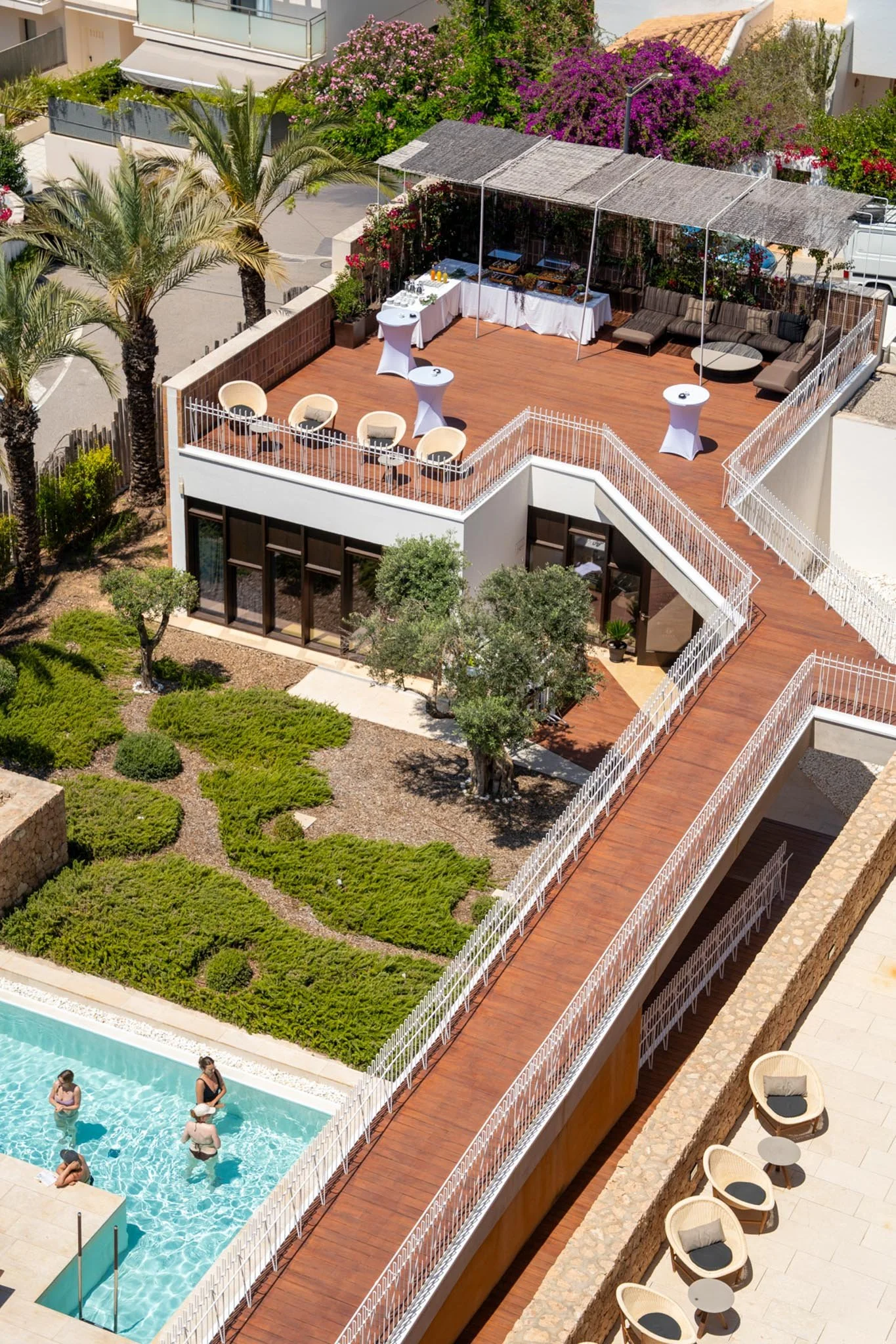 A rooftop swimming pool area with a small pool surrounded by a white fence, lounge chairs, and umbrellas. There's a wooden deck with tables and an outdoor seating area with a canopy. Palm trees and flowering bushes are visible around the area.