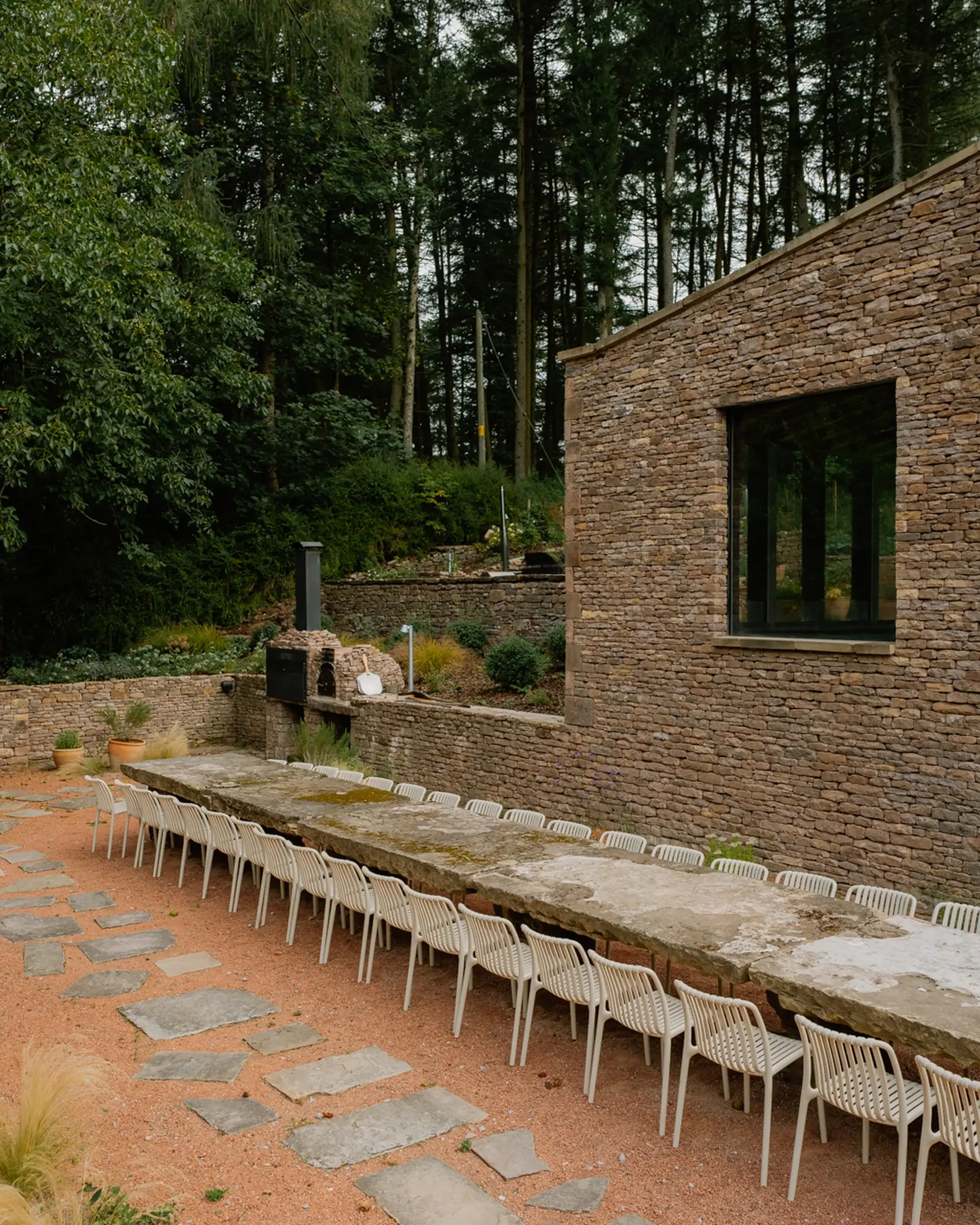 Outdoor patio area with a long stone table and white chairs, surrounded by a garden with potted plants, and a stone building in the background near a wooded area.