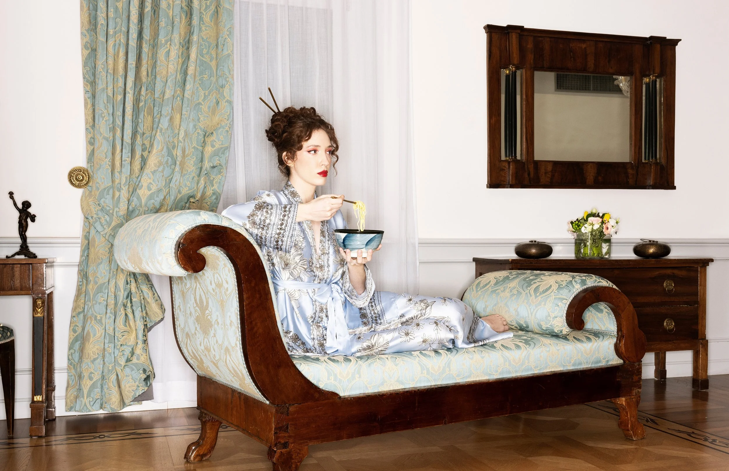 A woman with curly hair and red lipstick, dressed in a silk floral robe, is sitting on a vintage-style chaise lounge eating from a bowl of noodles with chopsticks. The room has elegant curtains, a mirror on the wall, and a small table with a floral a