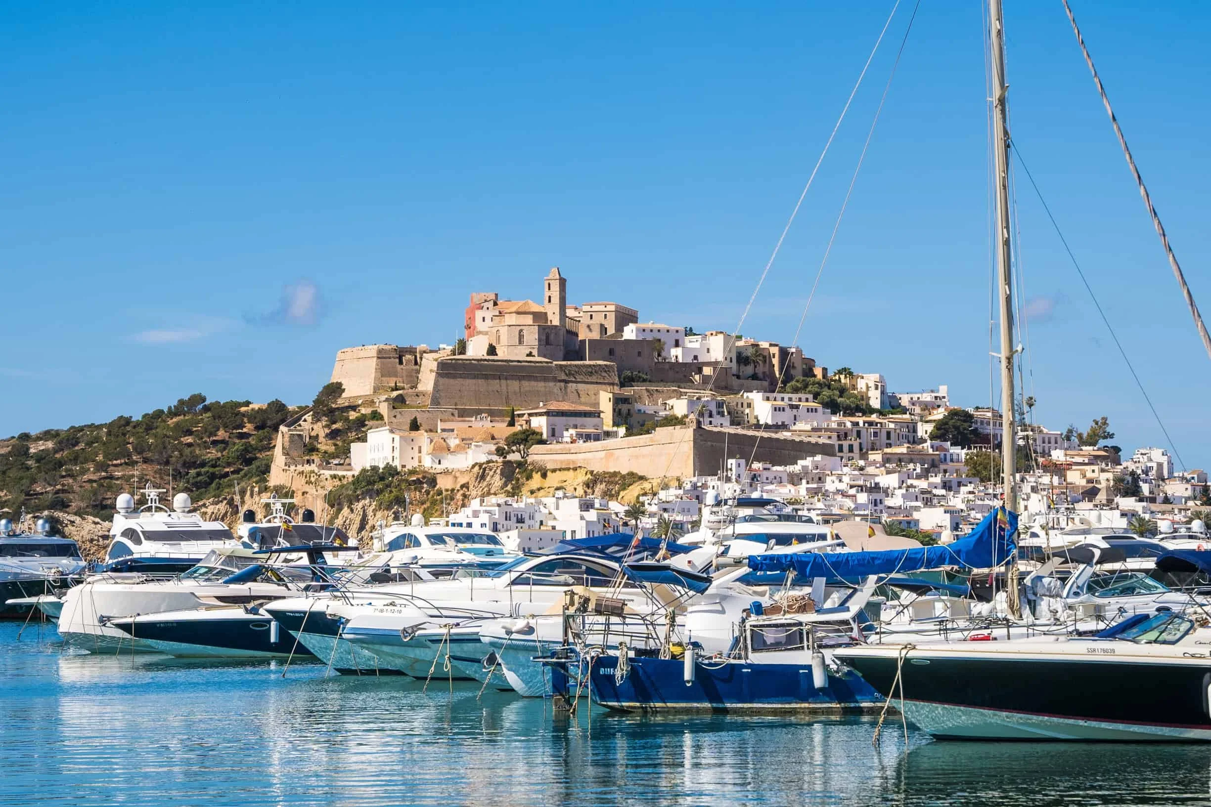 A marina filled with boats docked in calm water with a hillside town in the background, featuring historic buildings and a fortress on a hill.