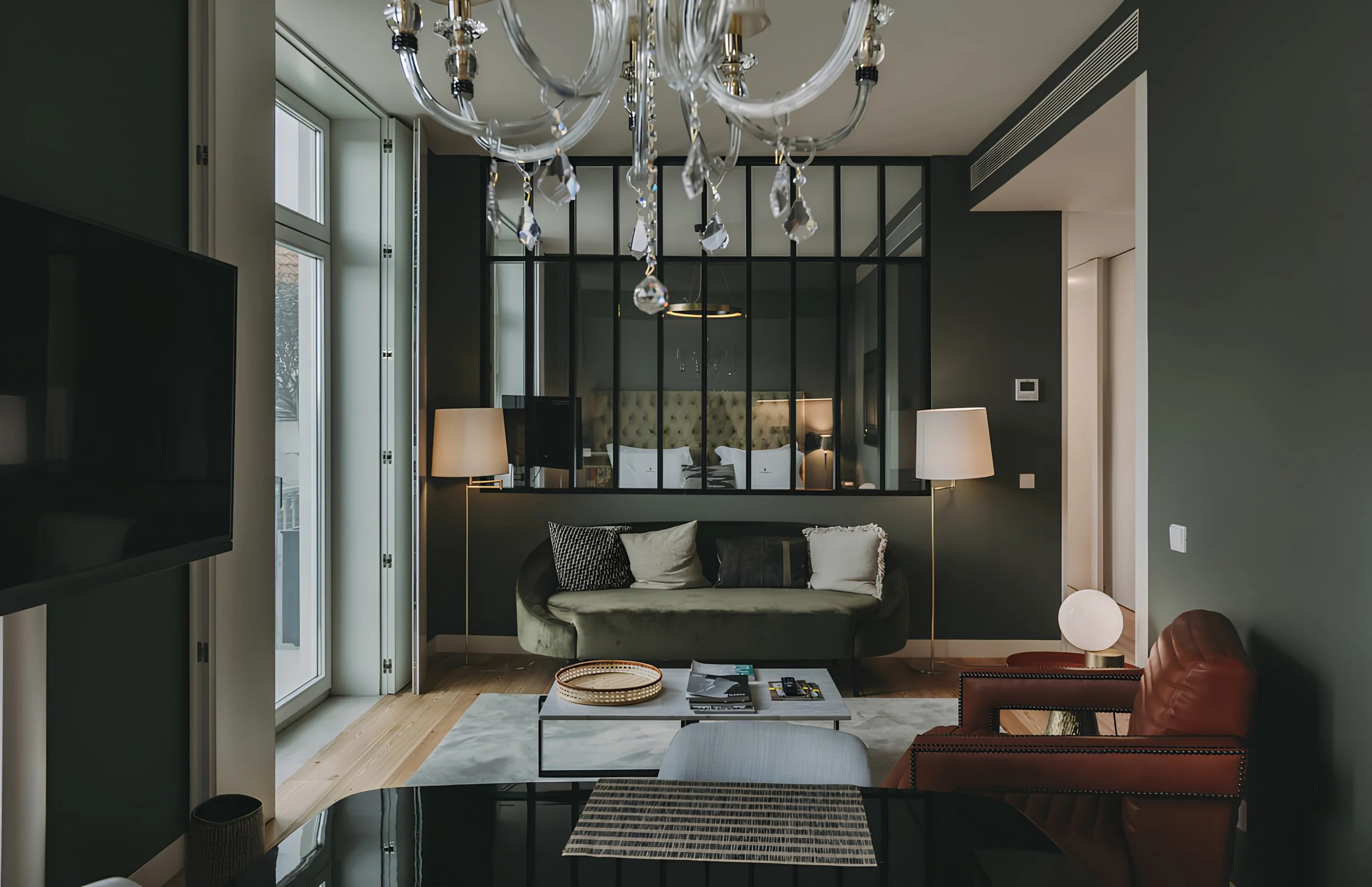 Living room with a dark wall, a light-colored sofa with pillows, a modern coffee table, a floor lamp, and a chandelier hanging from the ceiling. There is a window and a black-framed glass partition separating the living space from the bedroom with a 