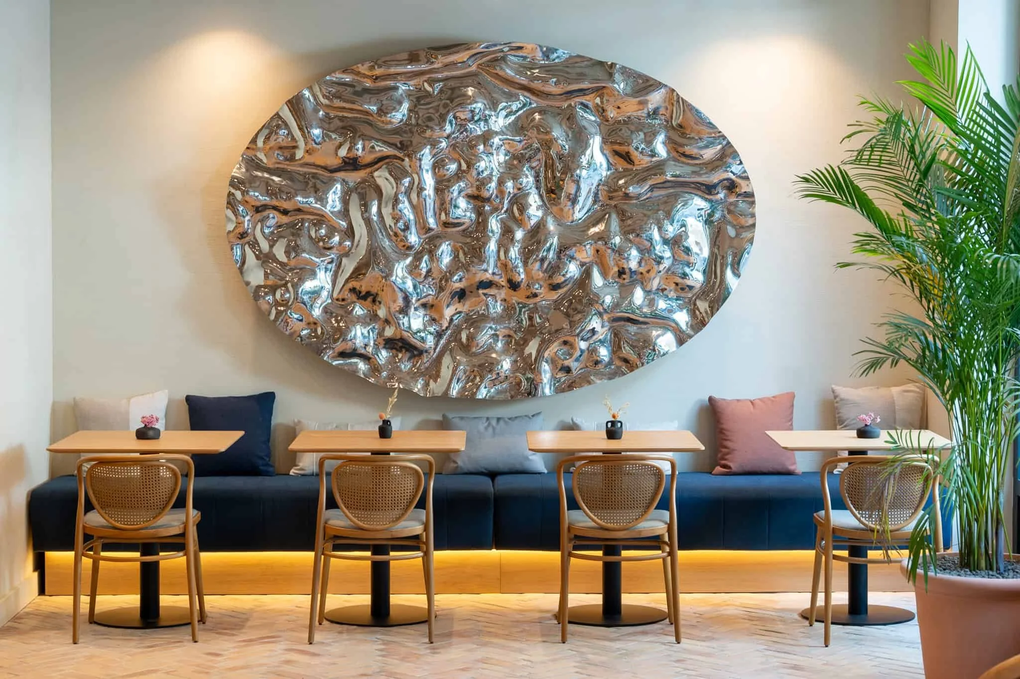 A modern dining area with four wooden chairs around a wooden table, small vases with pink flowers on each table, a large abstract metallic wall art piece, and lush green plants in the background.