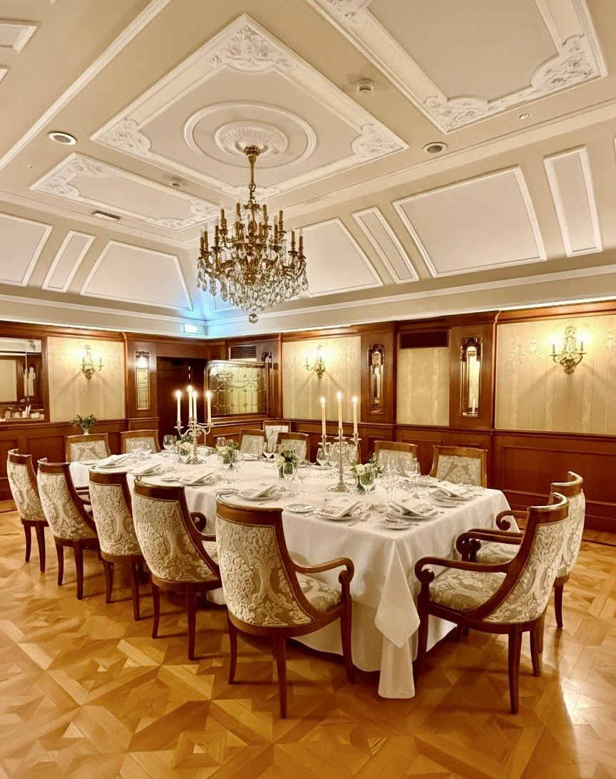 Elegant banquet room with a large rectangular table set for a formal dinner, surrounded by cushioned chairs with floral upholstery. The table is decorated with candlesticks, floral centerpieces, and fine china. The room features a chandelier, ornate ceiling details, wood-paneled walls, and wall sconces, with a parquet wood floor.