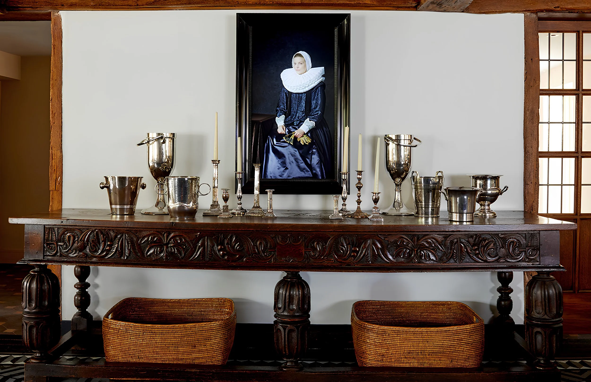 A wooden sideboard with intricate carvings holds silver candlesticks, vases, and bowls. Above it hangs a framed portrait of a woman in historical clothing with a ruffled collar, clutching gloves.