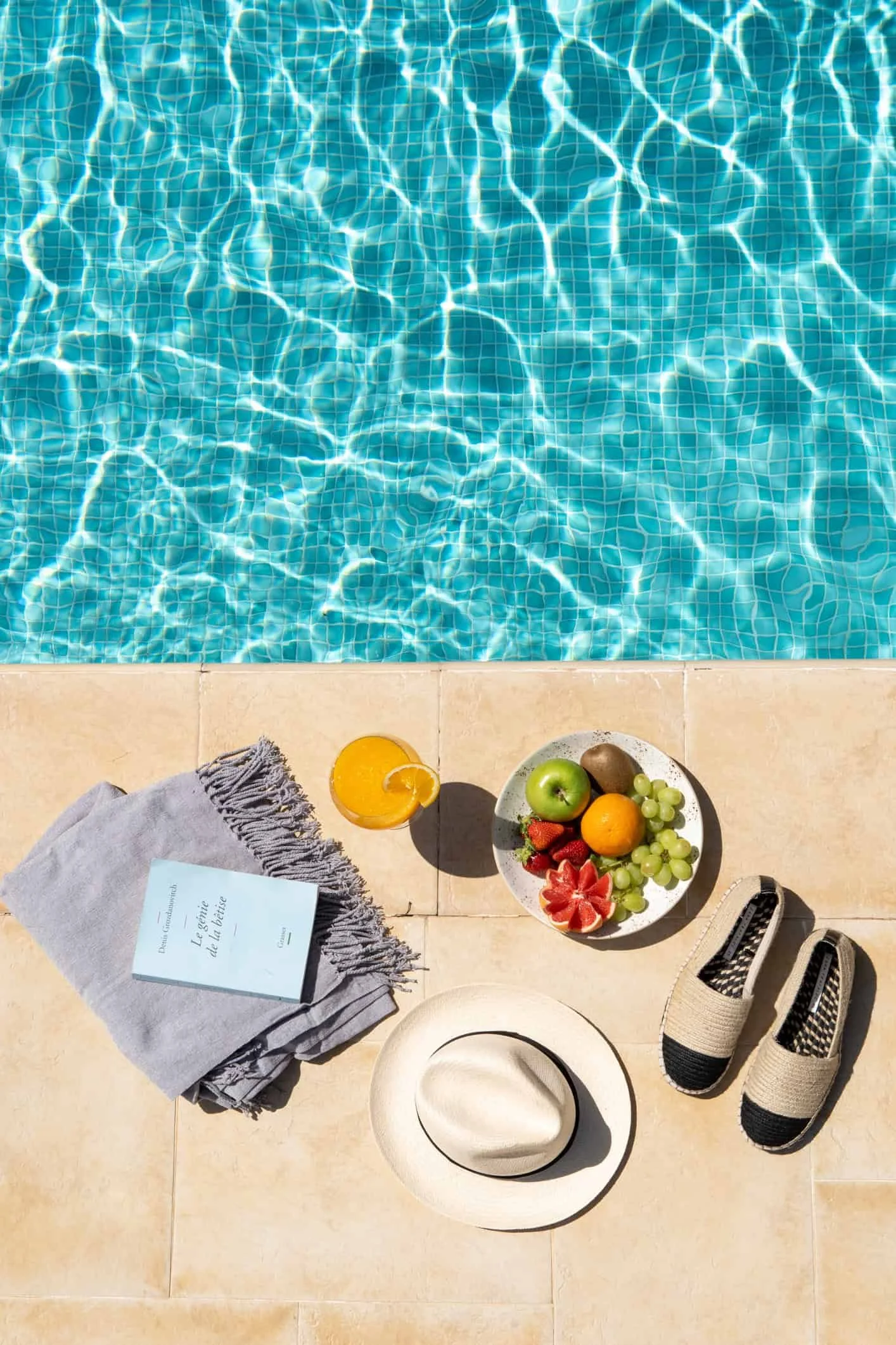 View of a swimming pool with blue water and tiled floor, nearby on the beige tiled ground are a gray towel, a white hat, a book, a bowl of assorted fruits, a pair of striped shoes, and a few other sun accessories.