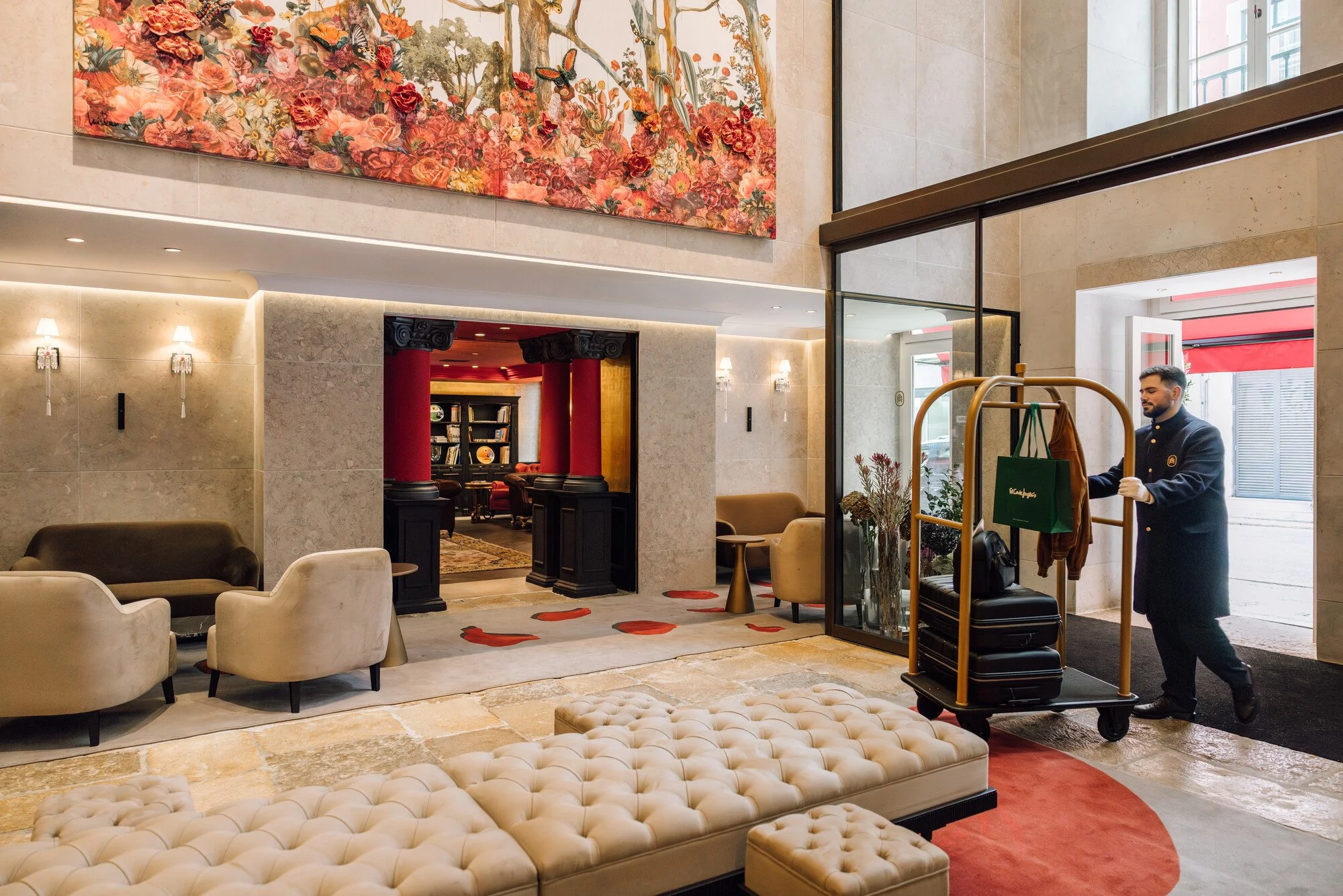The image shows a hotel lobby with modern decor. There are beige chairs and a tufted bench in the foreground. To the right, a hotel staff member is pushing a luggage cart with bags. The background reveals an interior space with a red and black color 