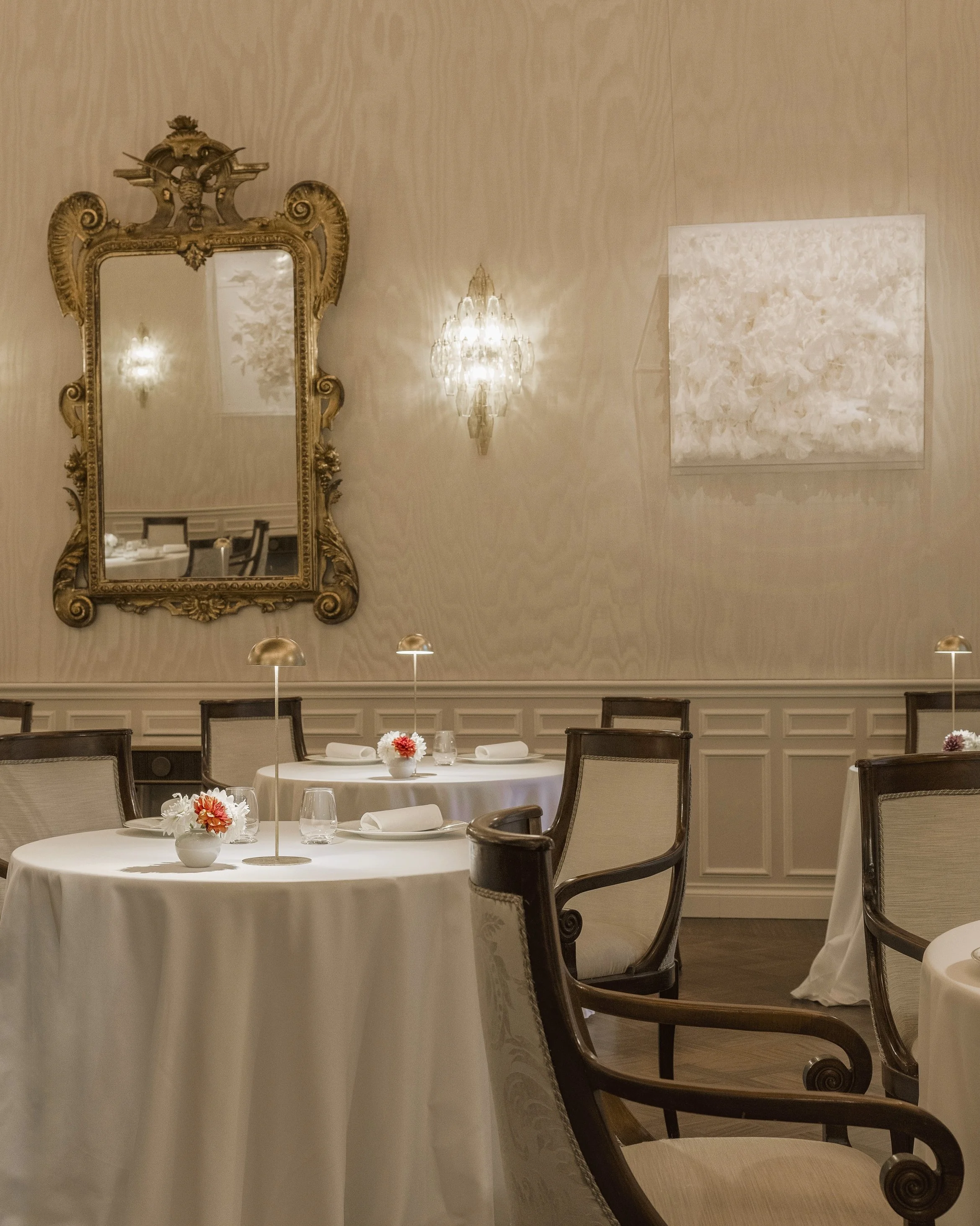 Elegant dining room with round tables covered in white tablecloths, decorated with flower centerpieces, surrounded by wooden chairs with beige upholstery, featuring a large ornate mirror, a crystal chandelier, and abstract artwork on the wall.