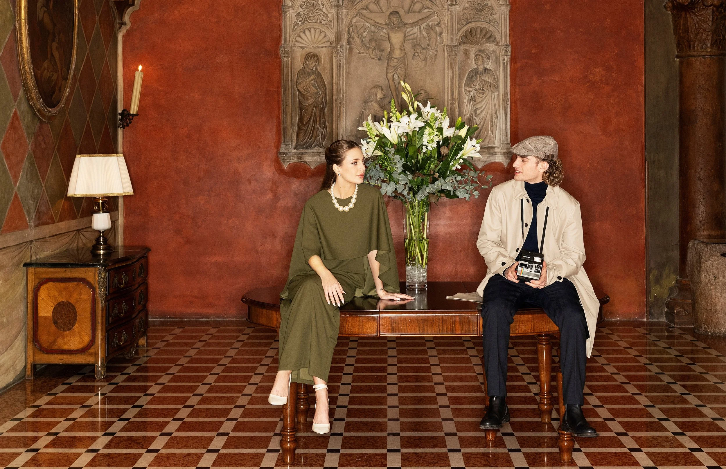 Two people sitting at a wooden table with a large floral arrangement of white lilies and greenery between them in a room with red and green painted walls, ornate wall carvings, and checkered tile flooring. The woman on the left wears an olive green o