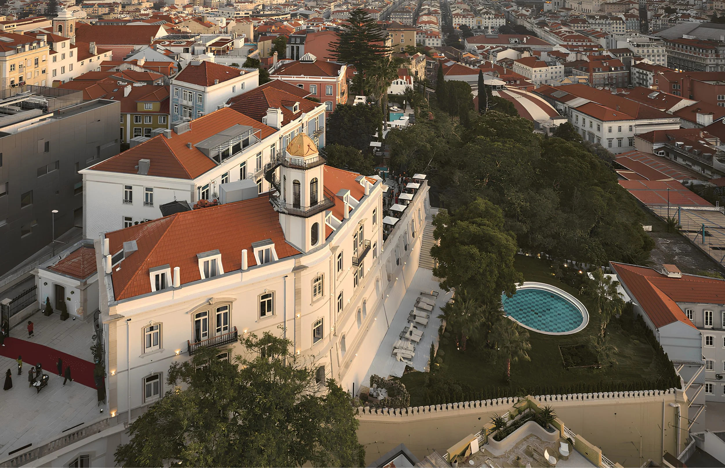 Aerial view of a city with many buildings featuring red-tiled roofs, green trees, and a swimming pool, likely at sunset.