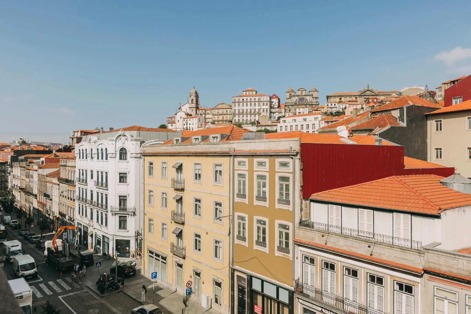 Cityscape with buildings featuring orange rooftops, and a hilltop with historic structures and churches under a clear blue sky.