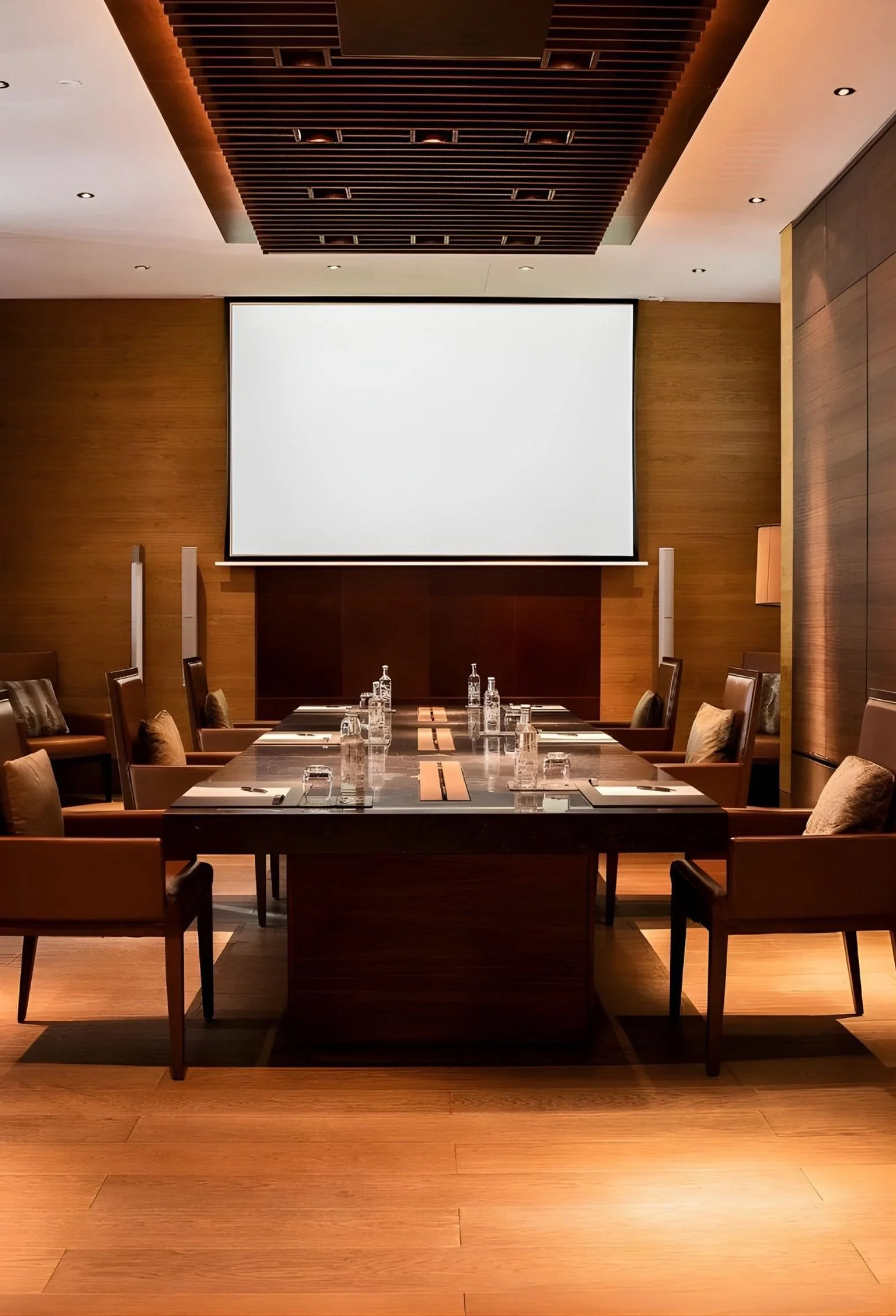 Conference room with a large blank white screen at the front, a long table set with water bottles, glasses, and notepads, surrounded by chairs with cushions, wood-paneled walls, and soft lighting.