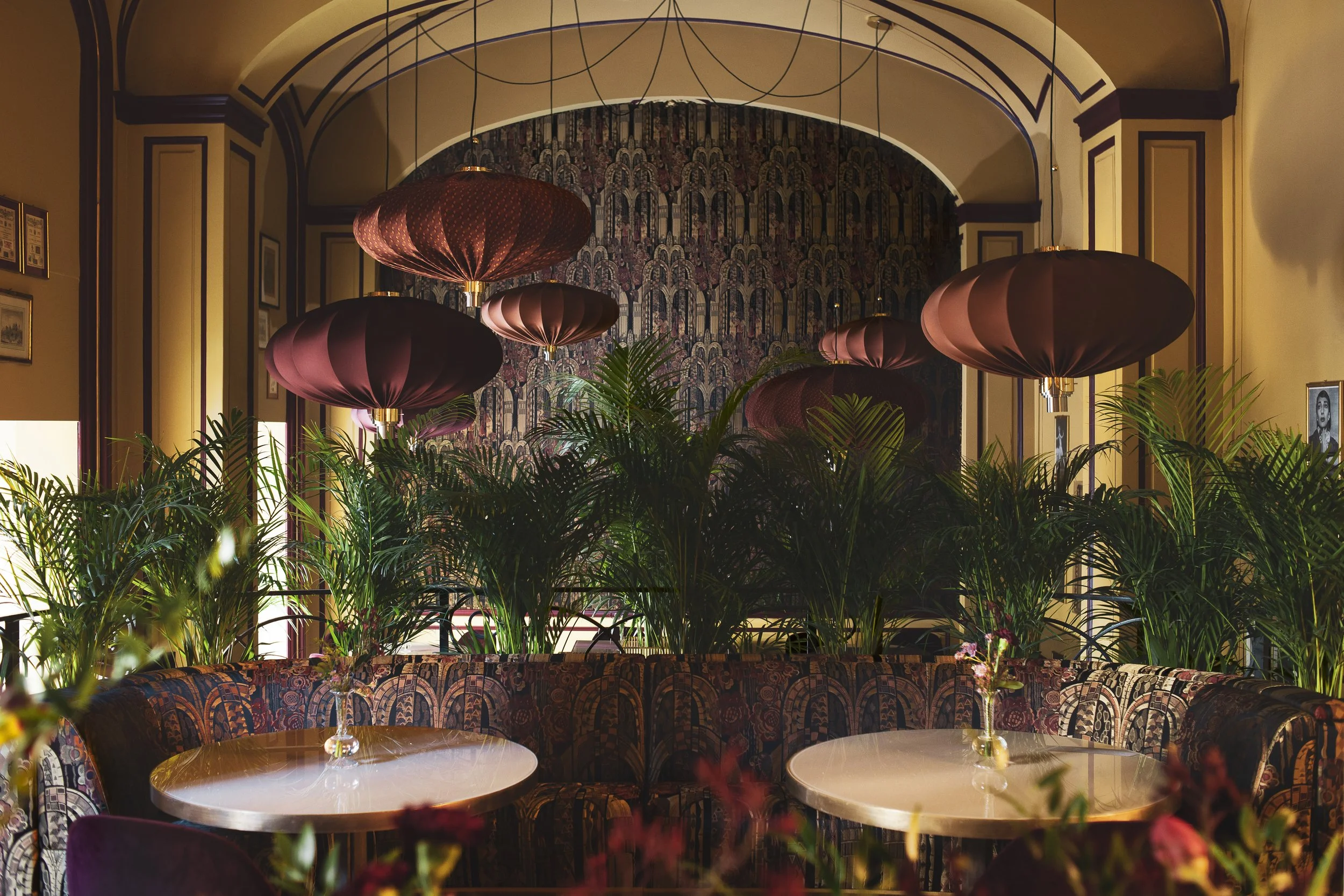 Elegant indoor space with patterned wallpaper, multiple pink and purple hanging lanterns, lush green plants, and a seating area with floral upholstery, illuminated by warm lighting.