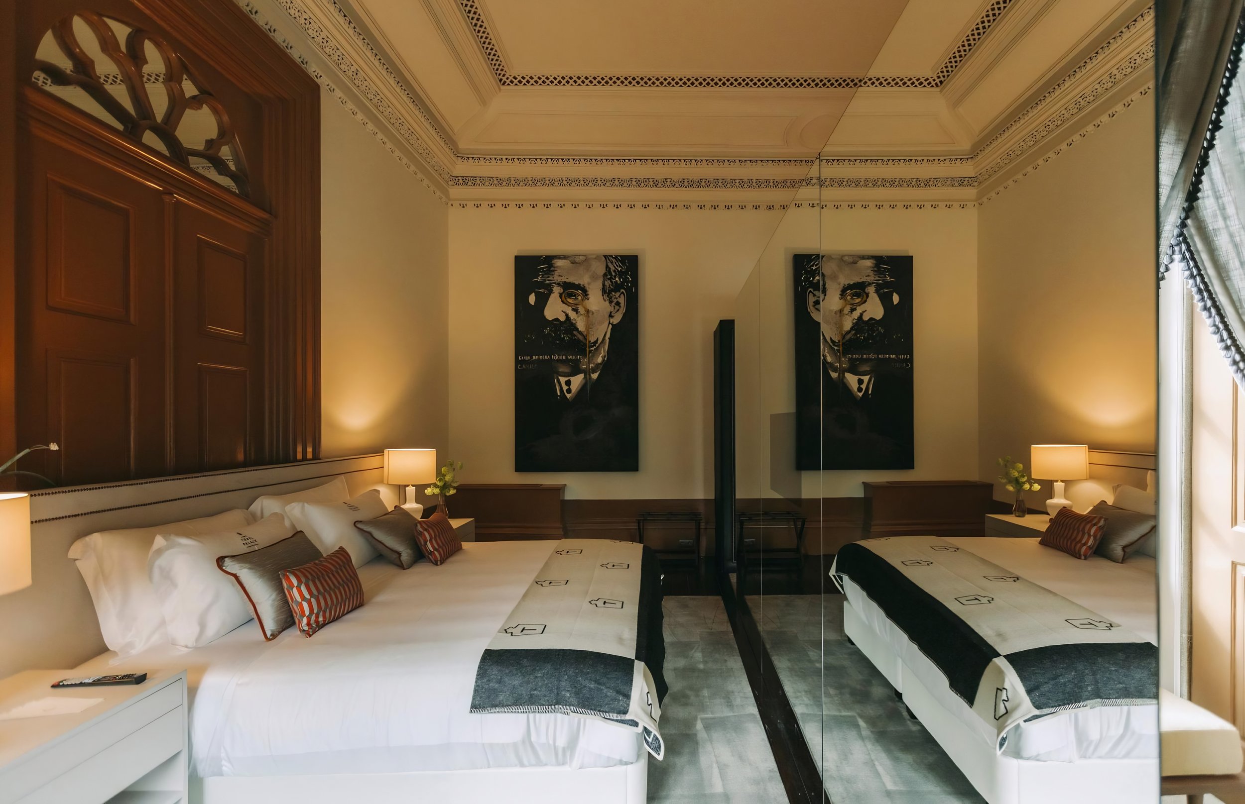 A hotel room with two beds, each with white linens and decorative pillows, and a mirrored wall with artwork of a man's face. There are nightstands with lamps on either side of the beds, and a window with curtains on the right.