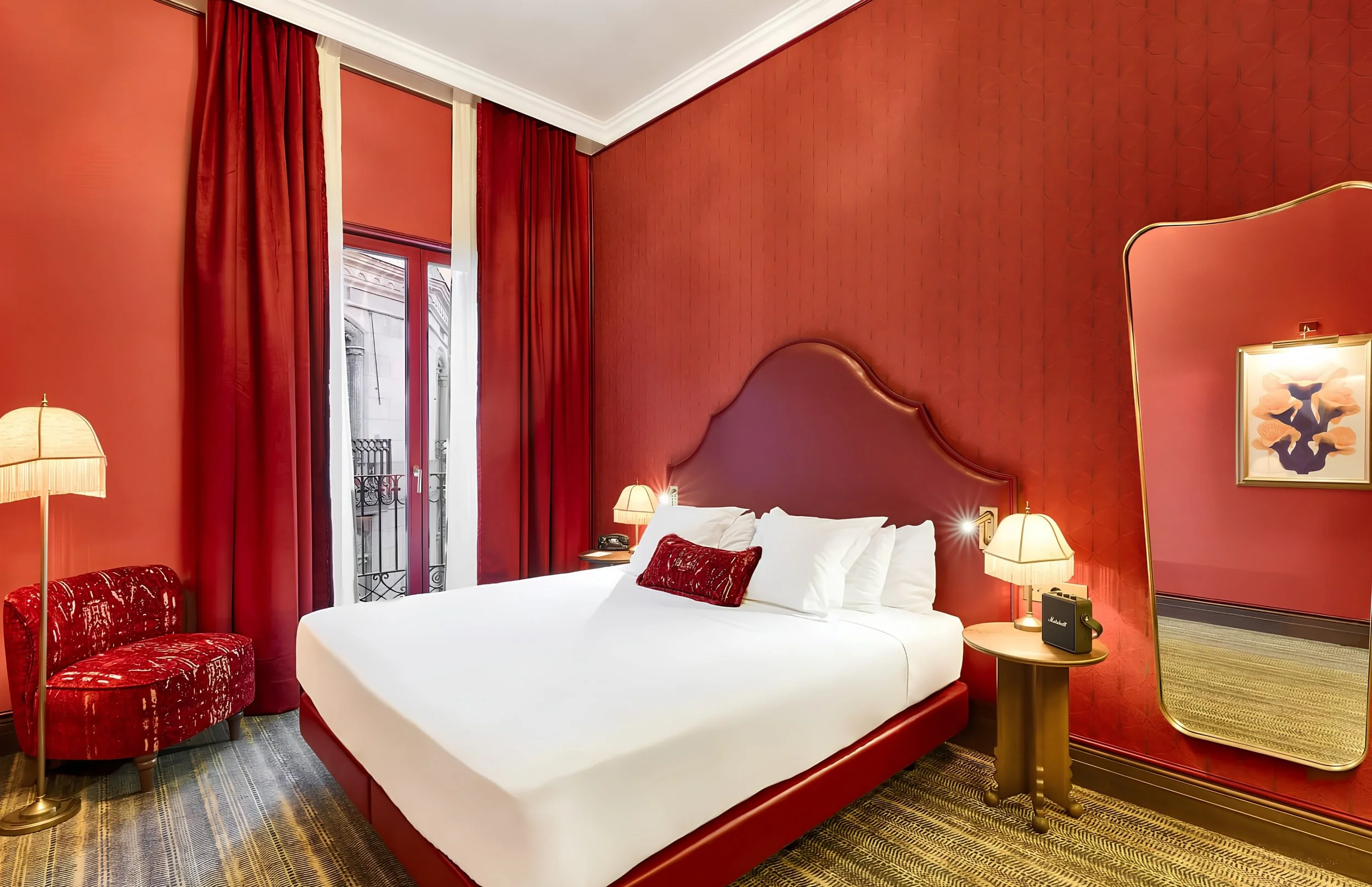 A luxurious hotel bedroom with a red theme. It features a large bed with white linens and red pillows, a padded red headboard, and matching red wall coverings and curtains. Beside the bed are two bedside tables with lamps, and a framed abstract flora
