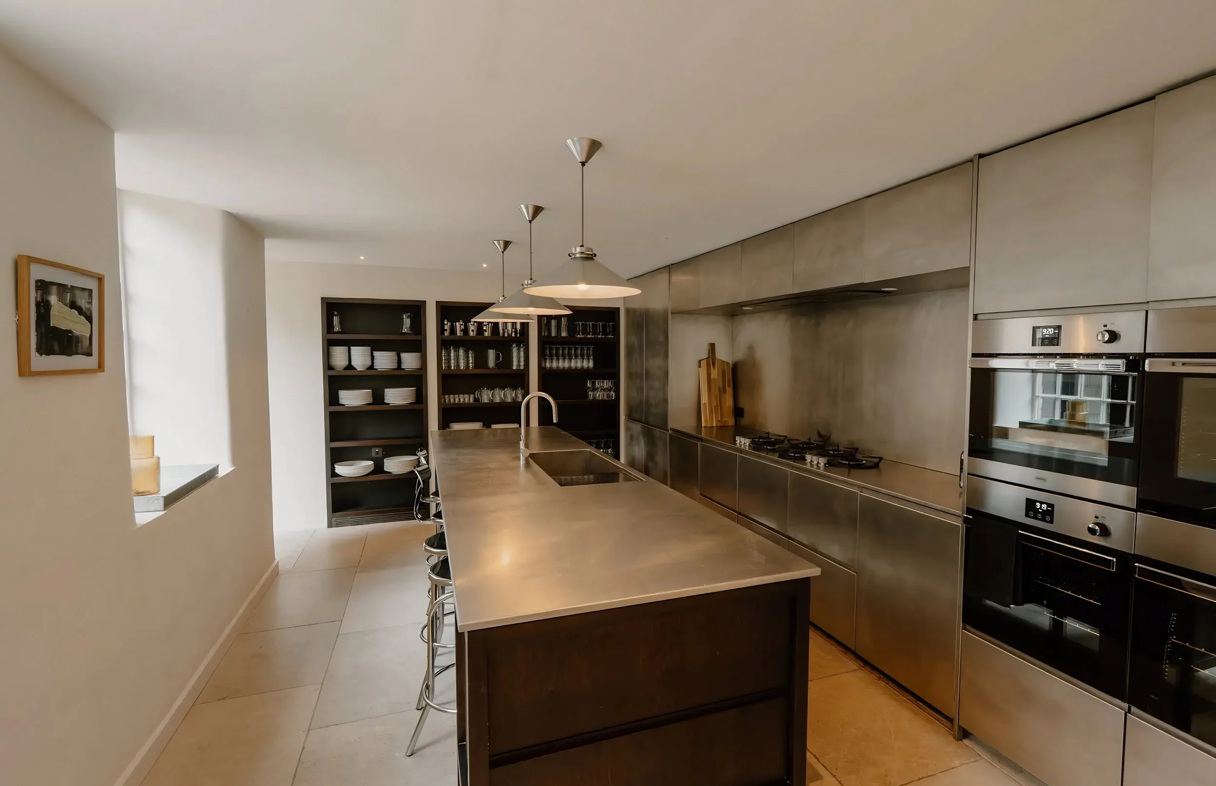 Modern kitchen with stainless steel appliances, open shelving with dishes, hanging pendant lights over a large island, and a built-in oven.