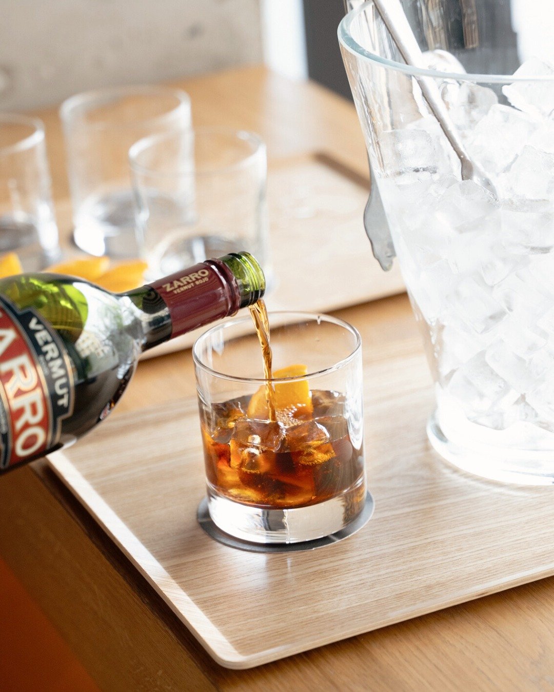 A glass of dark soda with ice and a lemon wedge, being poured from a bottle, on a wooden tray with empty glasses and a large ice bucket in the background.