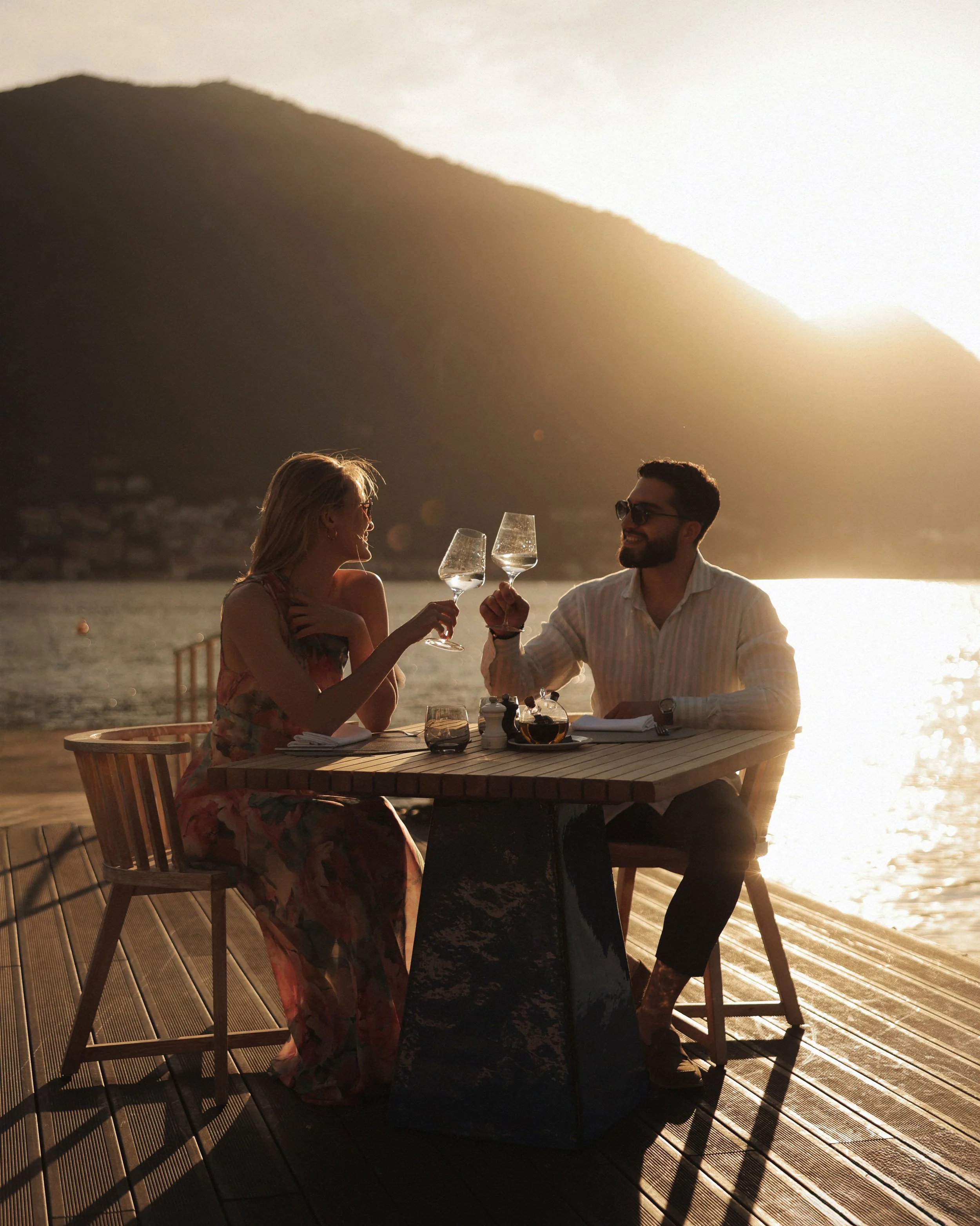A man and a woman enjoying drinks at a table on a wooden deck near the water during sunset, with mountains in the background.