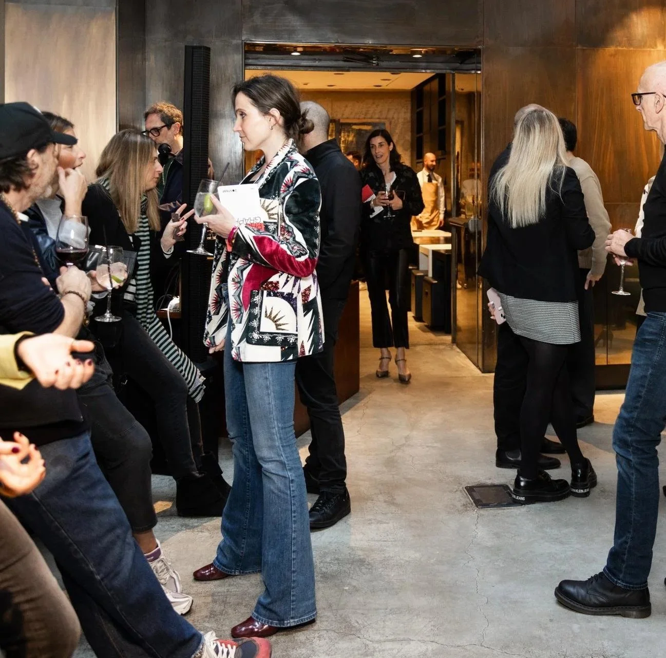 People mingling at a social gathering, some holding drinks, in a modern indoor setting with wooden and glass elements.
