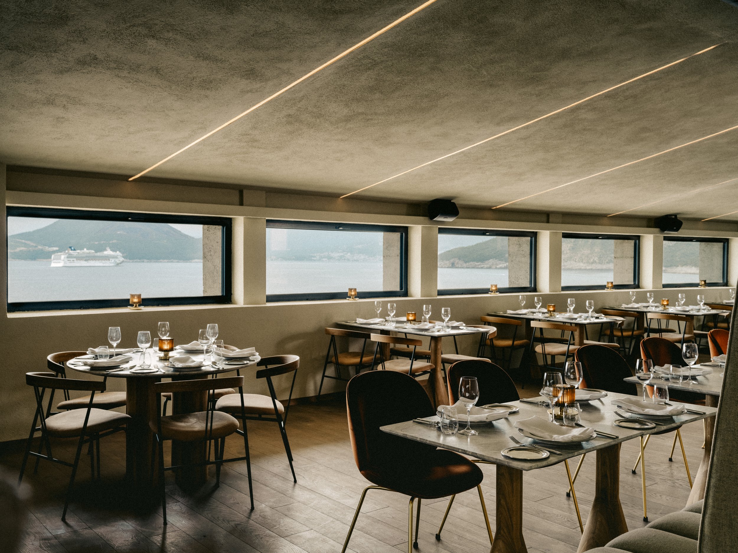 Modern restaurant dining area with round and square tables set with wine glasses, napkins, and candles. Large windows offer views of a harbor with a cruise ship and mountains in the distance.