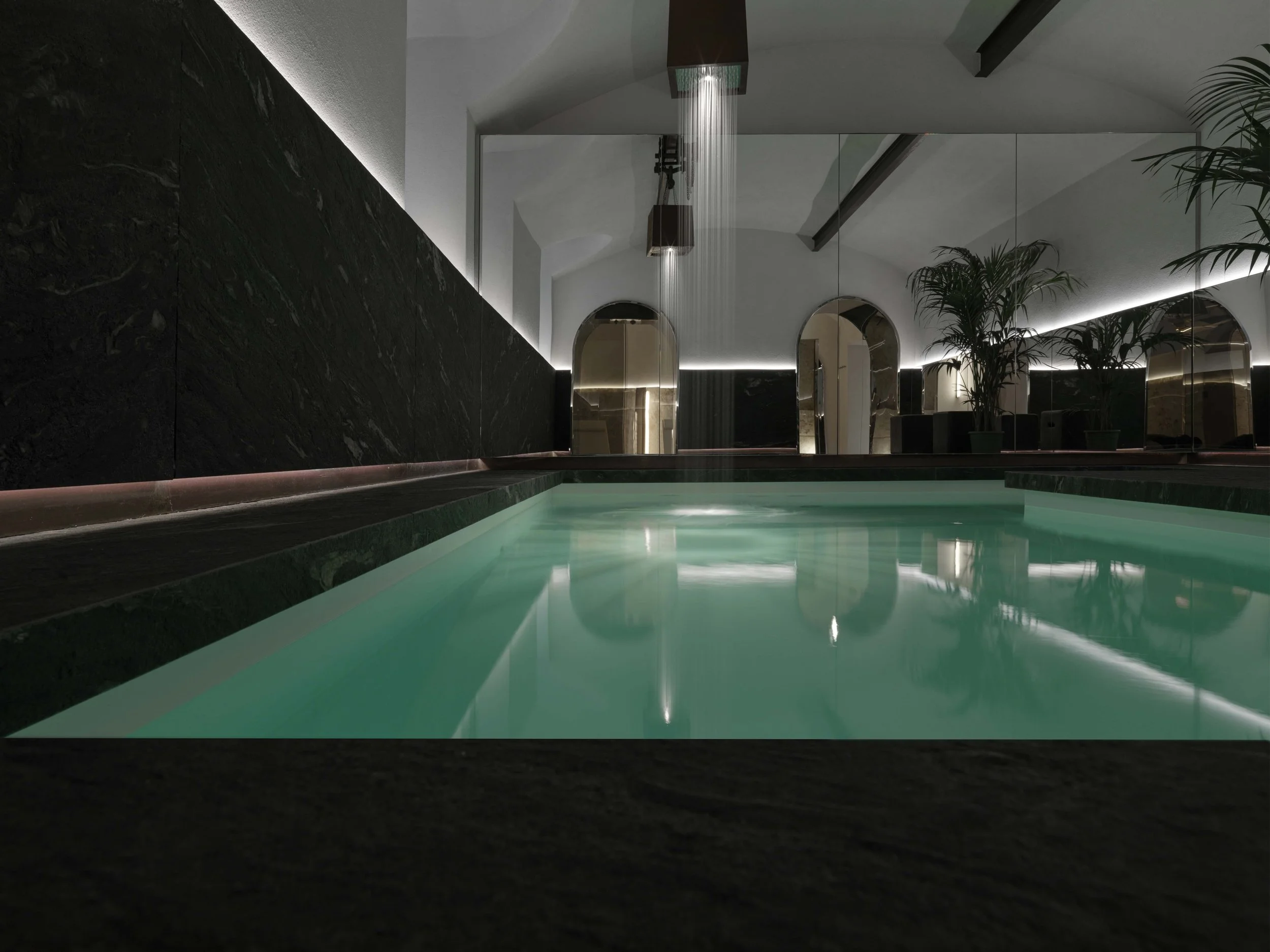 Indoor swimming pool with modern decor, dark marble walls, a water feature with rain shower, large mirrors, potted plants, and ambient lighting.