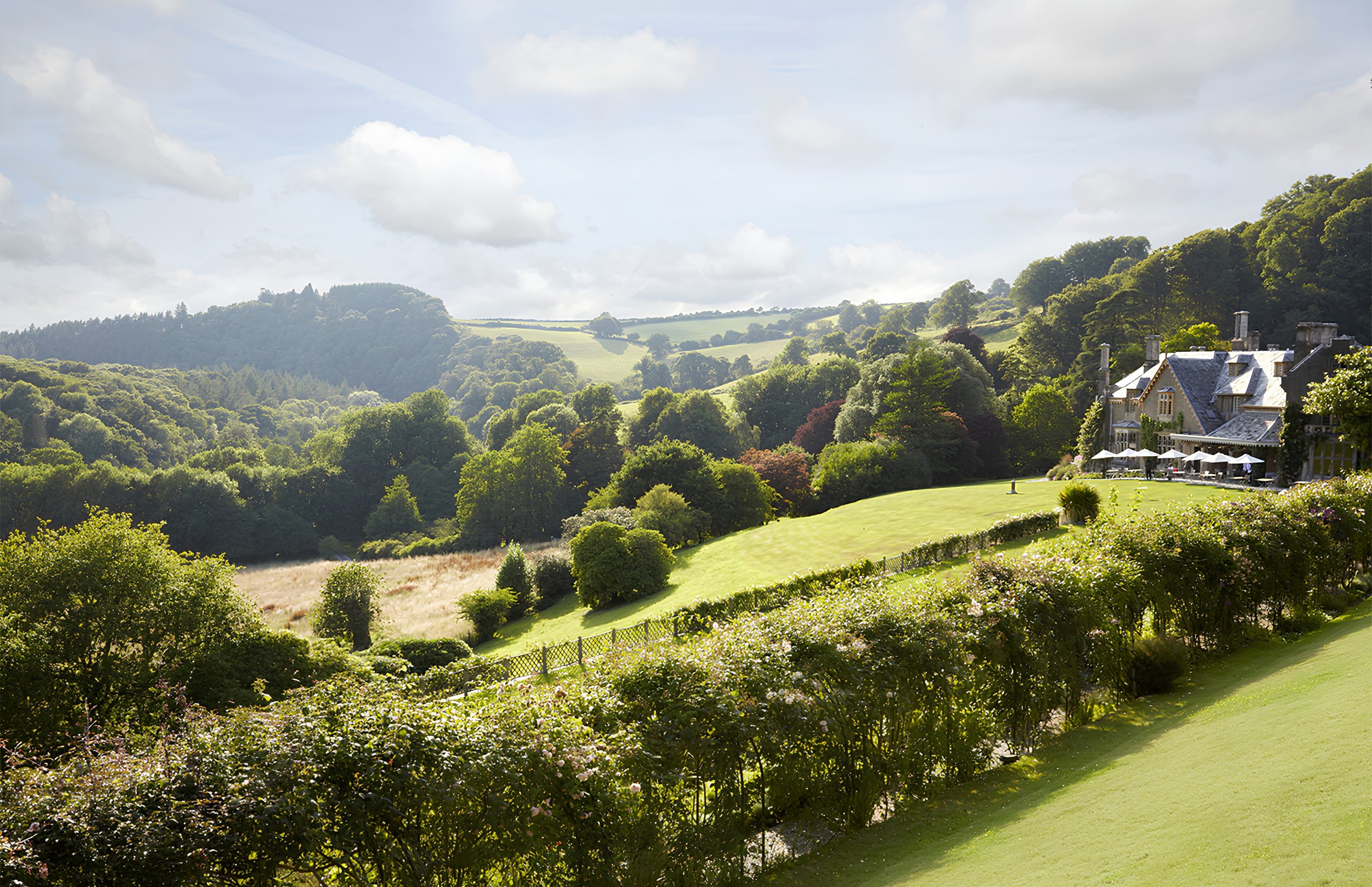A lush green landscape with rolling hills, trees, bushes, and a large estate with outdoor seating and umbrellas.