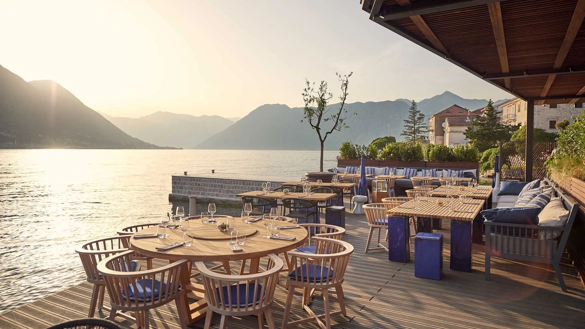 An outdoor dining area on a waterfront patio with a lake, mountains, and a sunset in the background. There are wooden tables set with glassware and cutlery, surrounded by chairs with blue cushions, and cushioned seating along the edge.