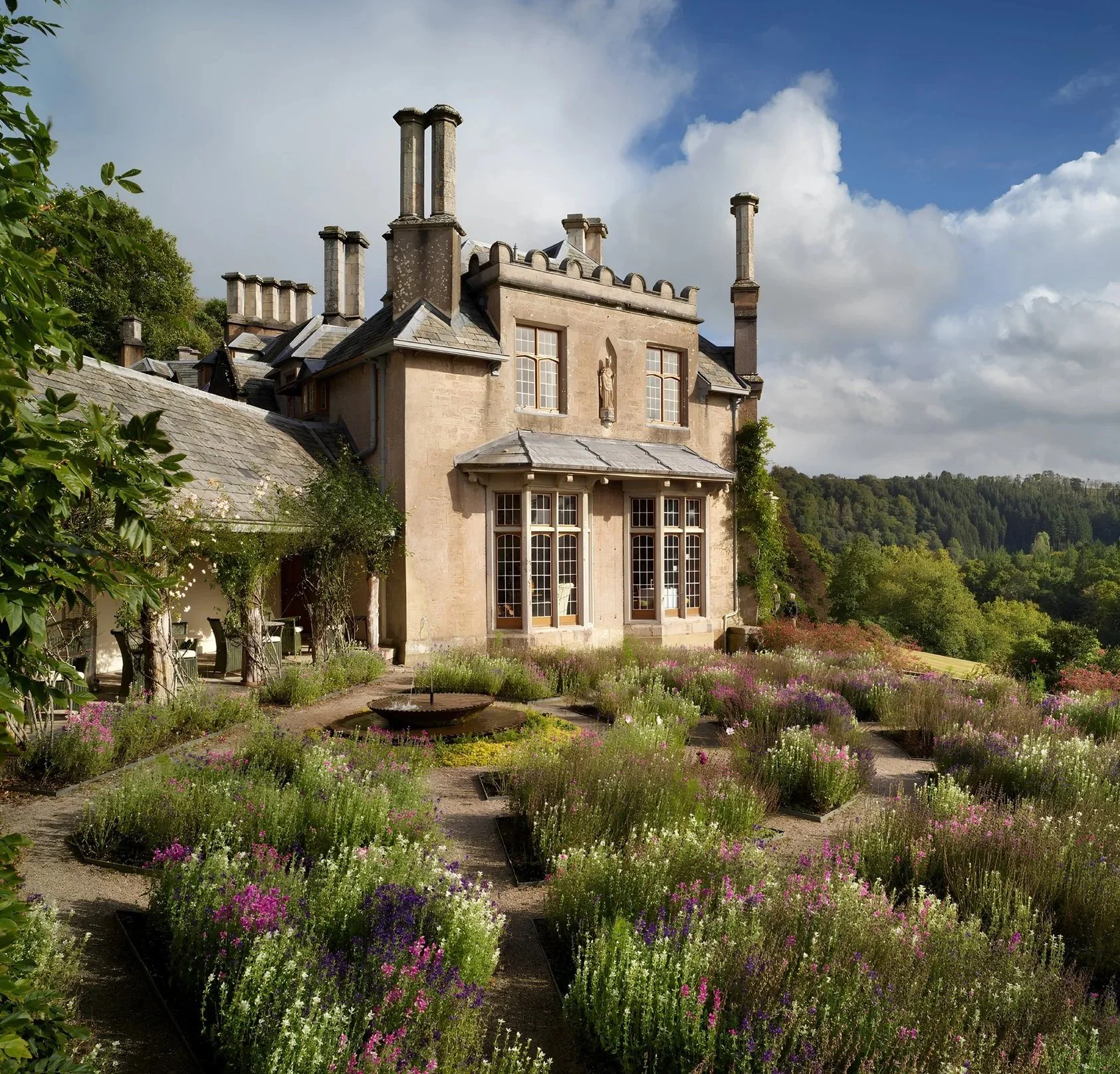 A large historic stone house with multiple chimneys, surrounded by a colorful garden with flowers, under a partly cloudy sky.