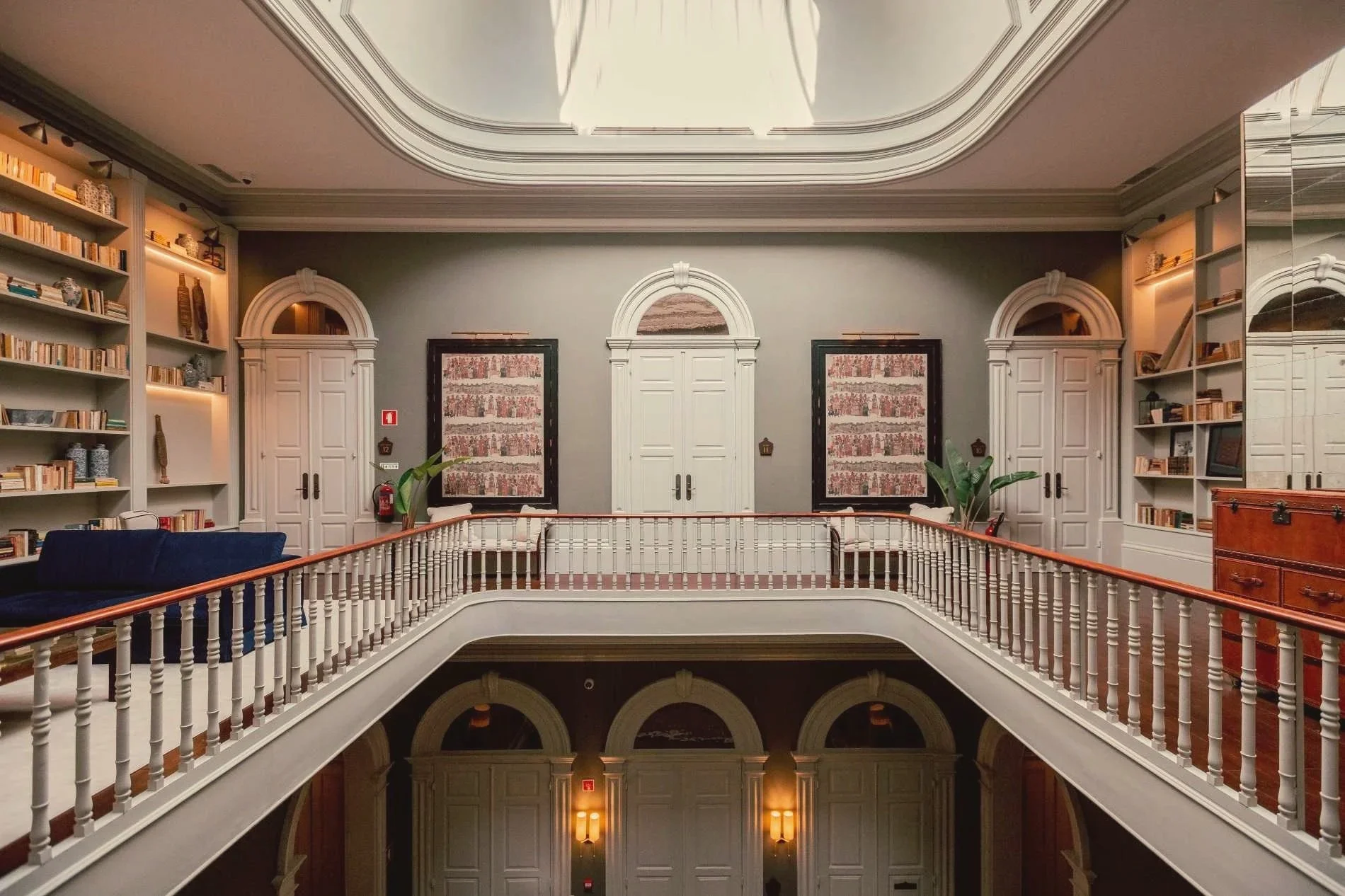 Interior view of a two-story building with a balcony, ornate ceiling, bookshelves filled with books and decorative items, framed artwork, plants, and wooden furniture.