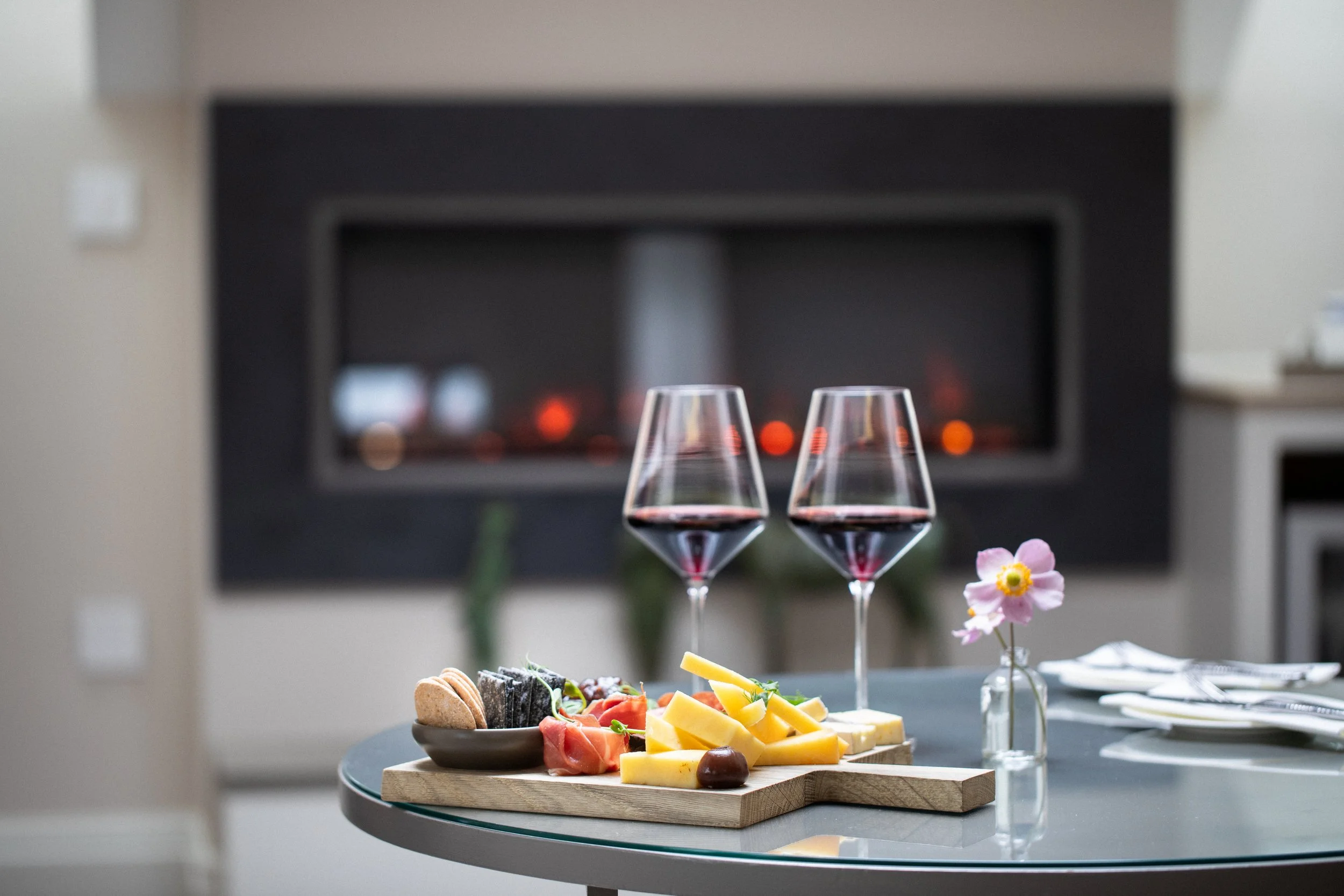 Elegant table setting with wine glasses, cheese, grapes, and a charcuterie platter in front of a modern fireplace.